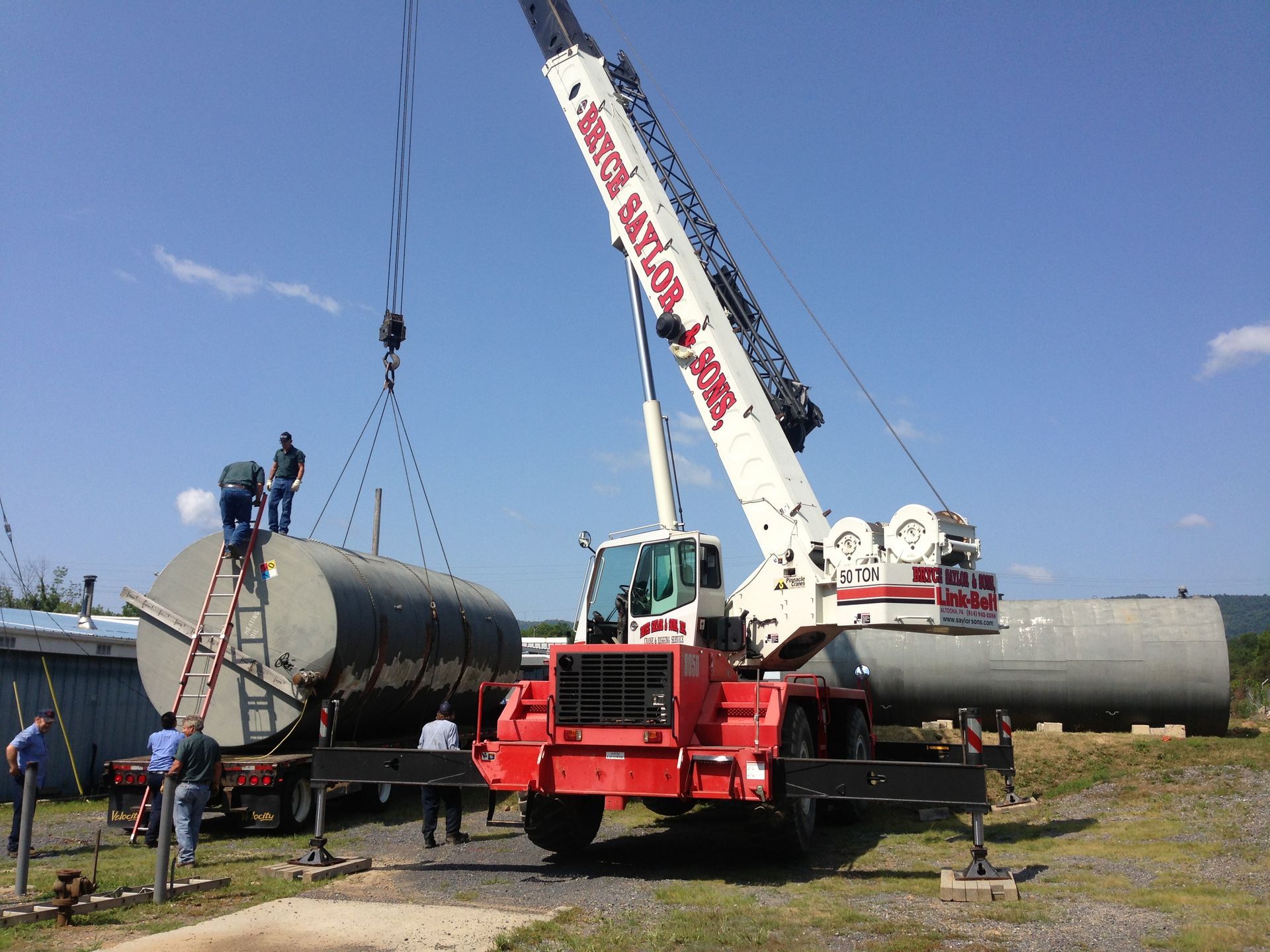 Crane lifting a large horizontal tank at an outdoor work site under a blue sky