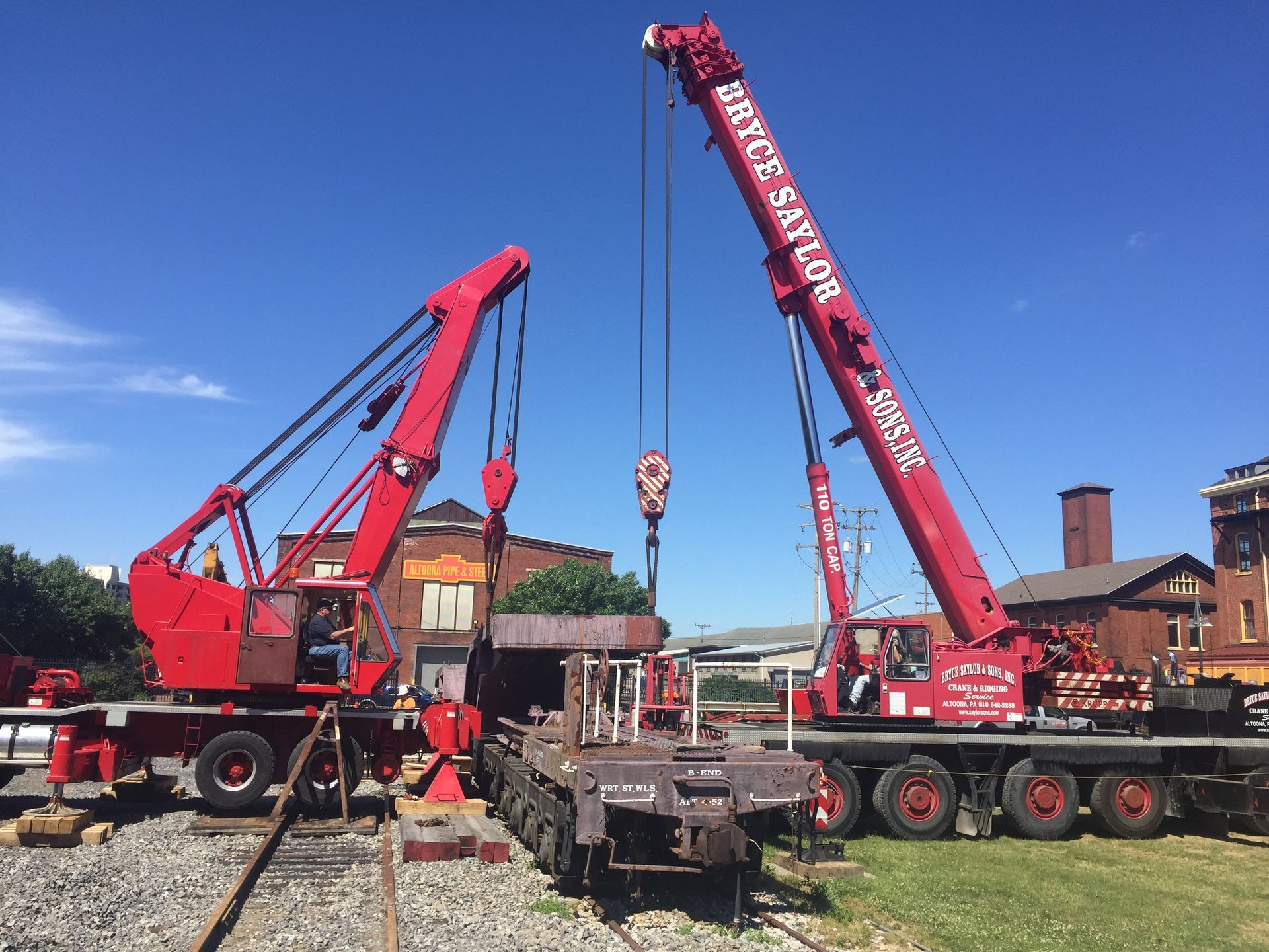 Two red mobile cranes lifting a heavy object in an industrial yard under a blue sky