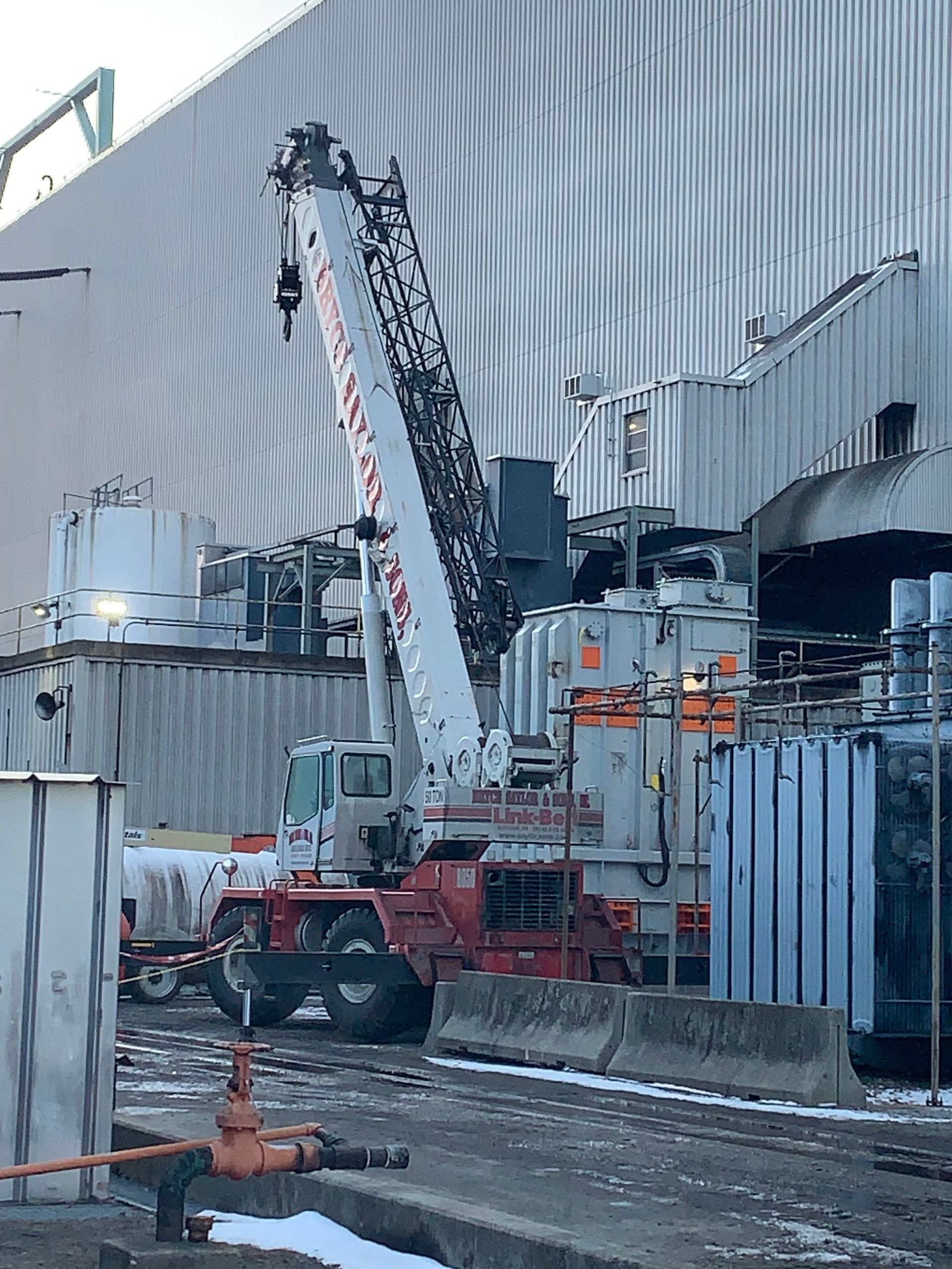 Red and white crane truck lifting beside a gray industrial building and fence.