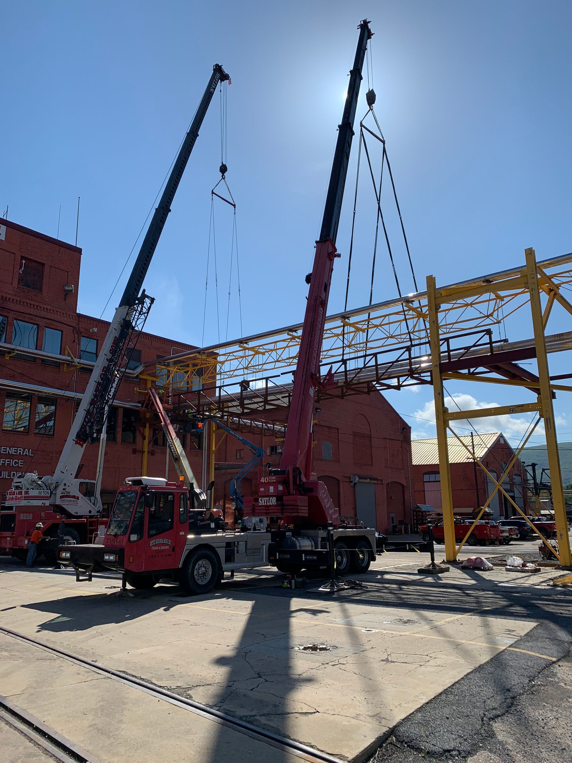 Two red cranes lifting steel beams at a construction site beside brick buildings and a yellow frame.