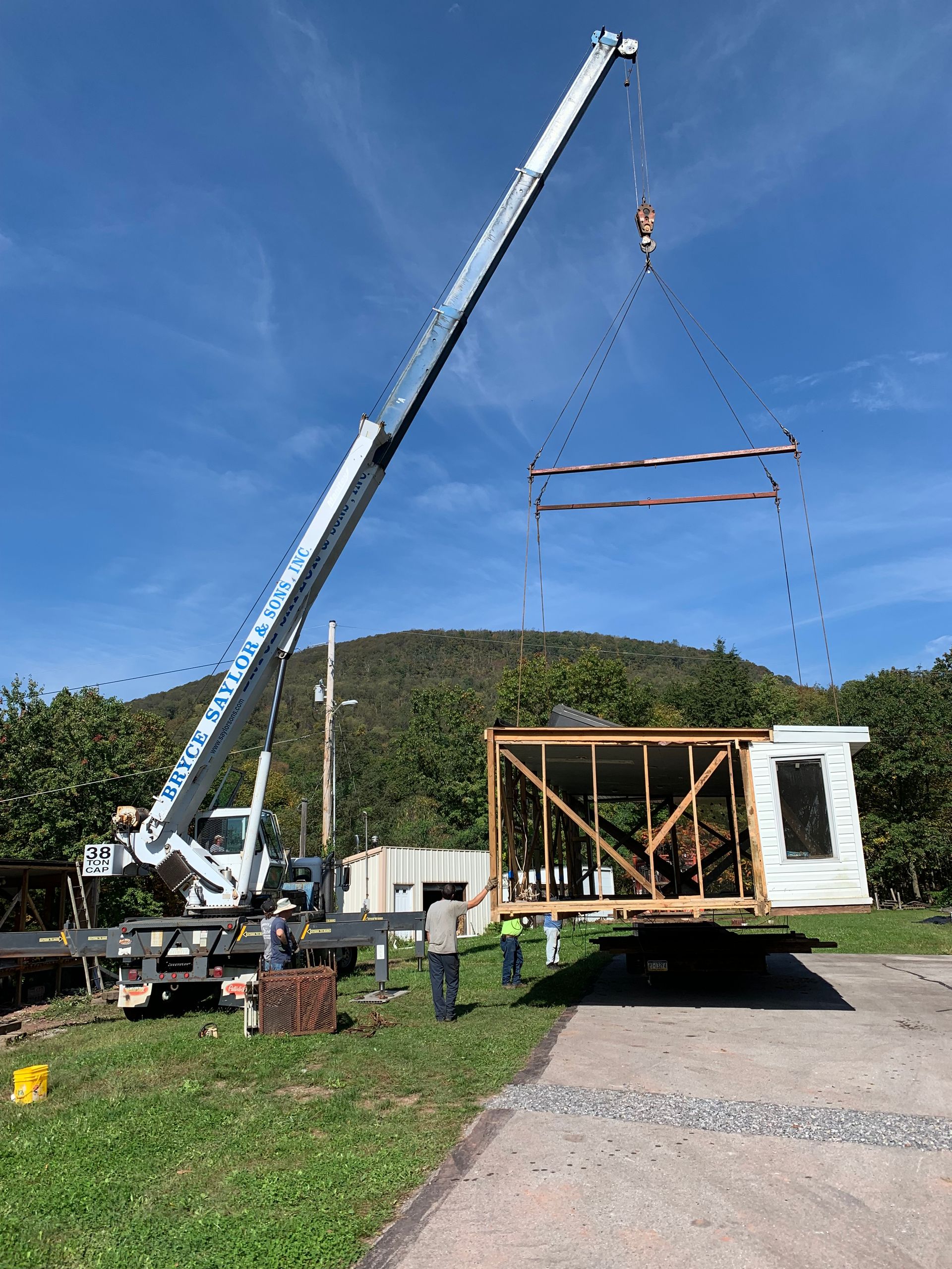 Crane lifting a wooden building frame beside a mobile home in a rural field under a blue sky