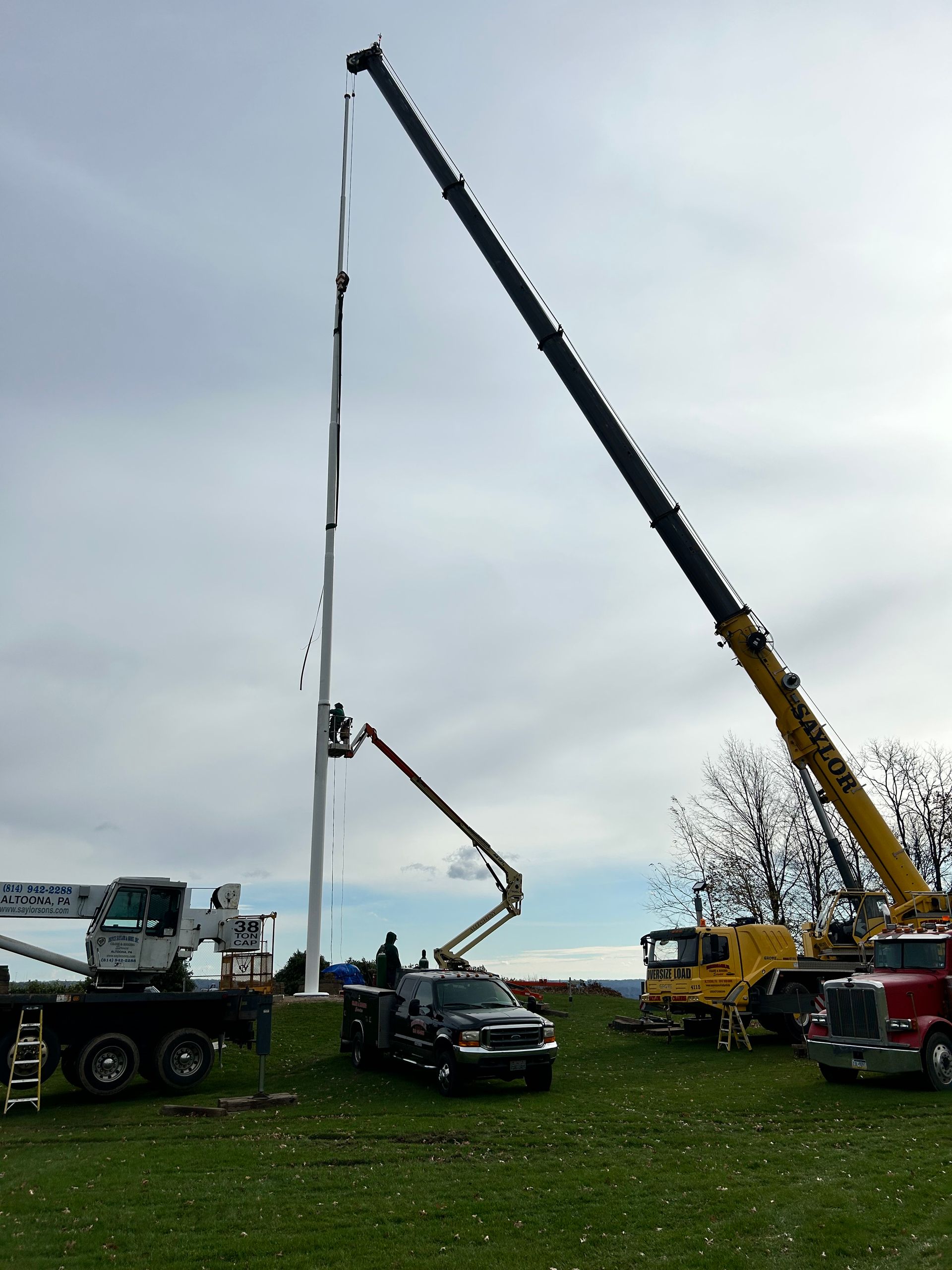 Two cranes lifting a tall pole on a grassy field under a cloudy sky