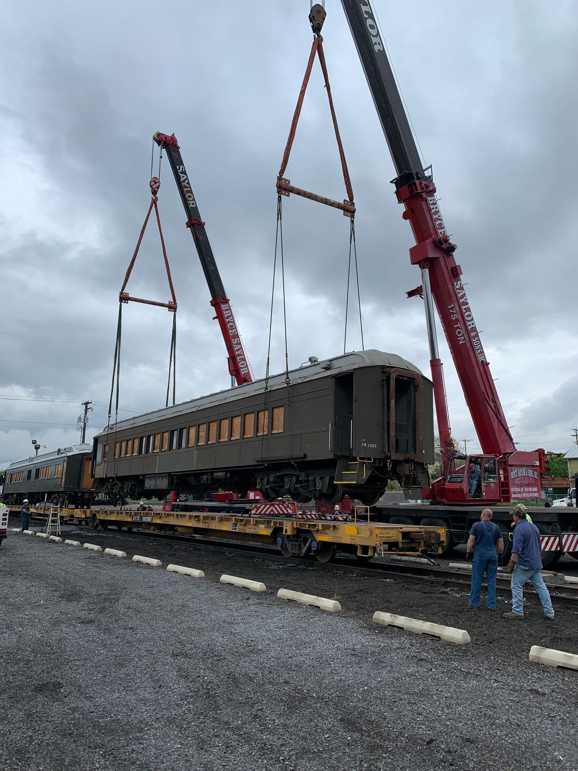 Heavy cranes lifting a large railcar on tracks in a gravel yard under a cloudy sky