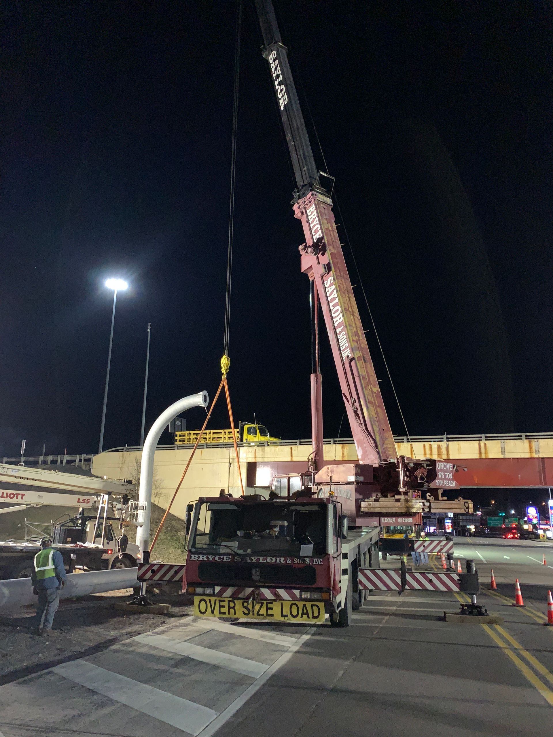 Night roadwork scene with a crane lifting equipment beside a highway overpass under streetlights.