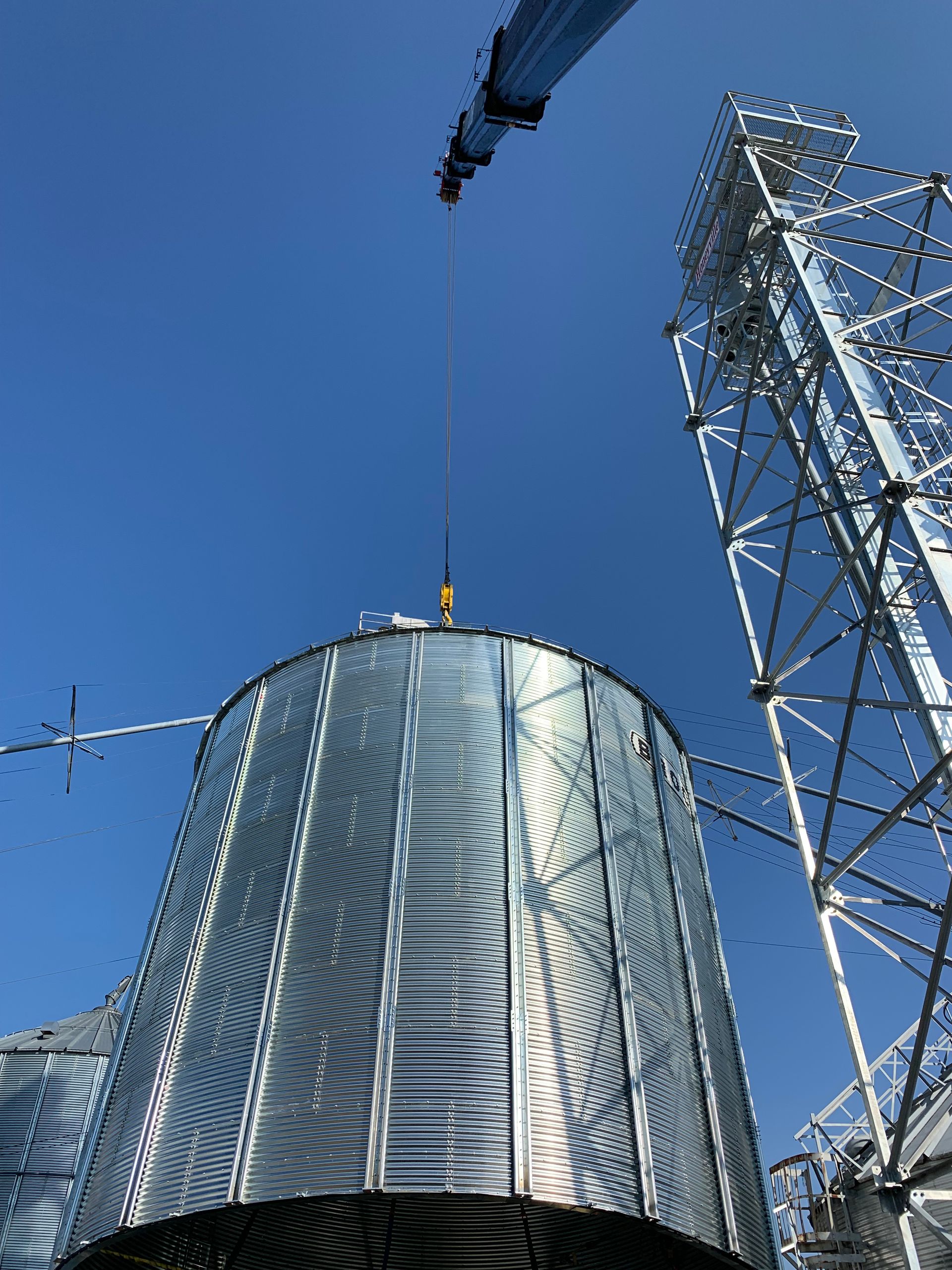 Crane lifting a shiny metal silo beside a tall lattice tower under a clear blue sky