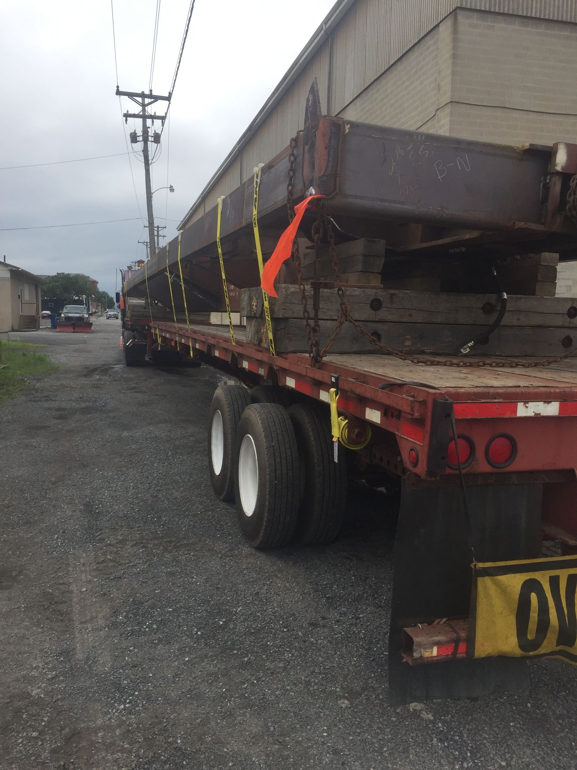 Flatbed trailer hauling a long metal structure on a gravel lot under cloudy skies.