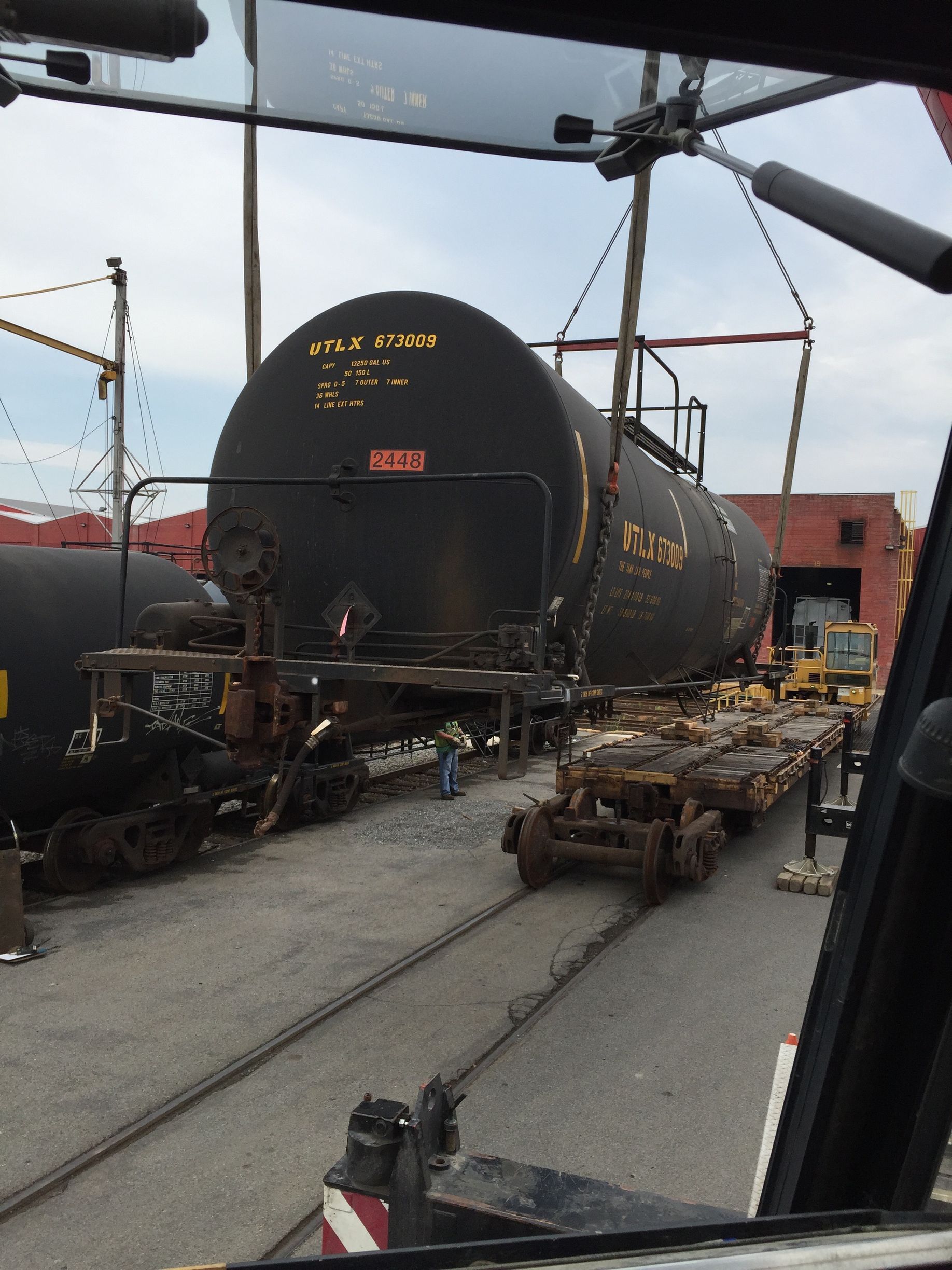 Large black cylindrical tank car on rail yard tracks, with maintenance equipment around it under an overhead structure