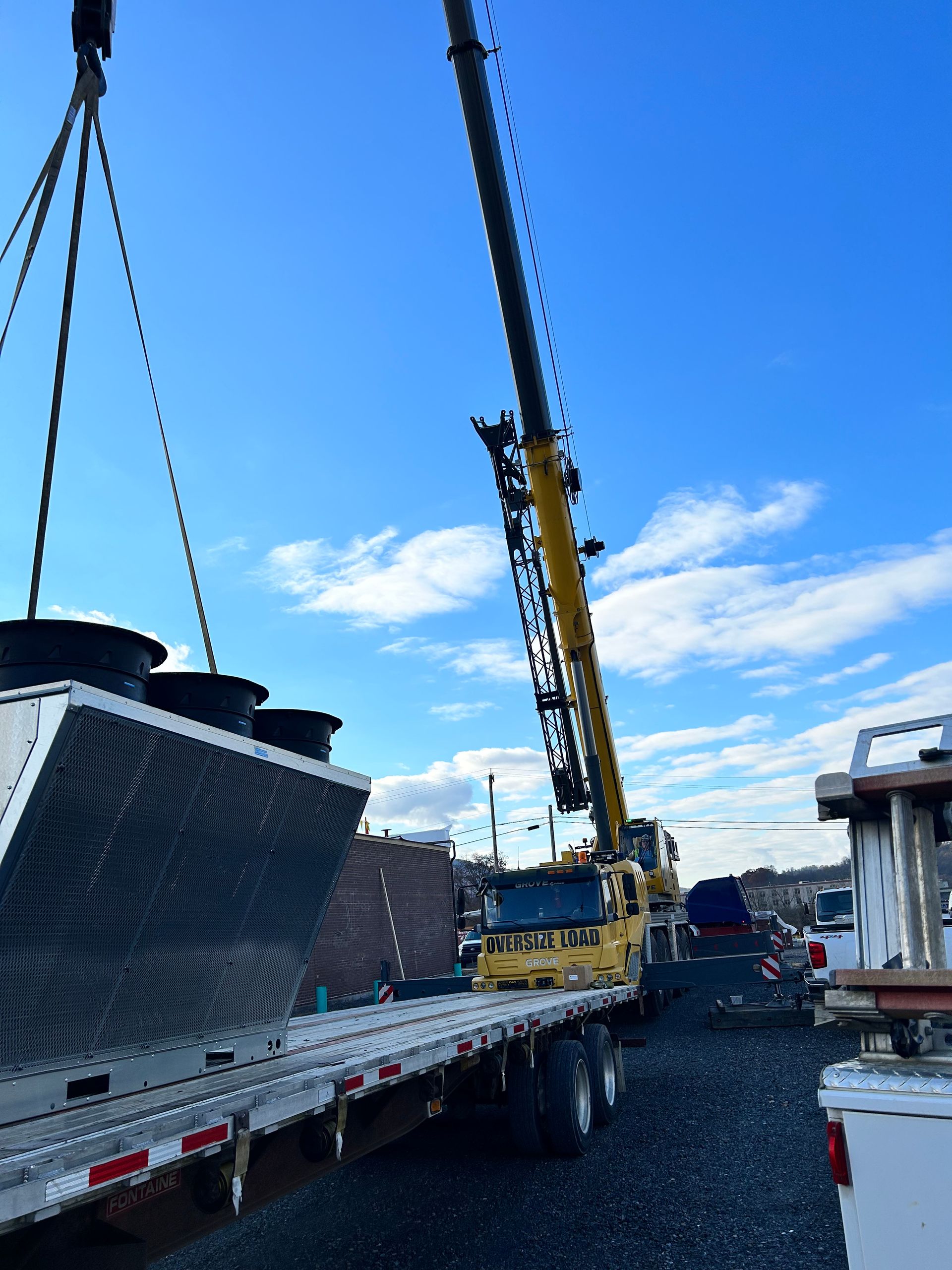 Flatbed truck carrying a large industrial unit, lifted by a crane in a port or construction yard.