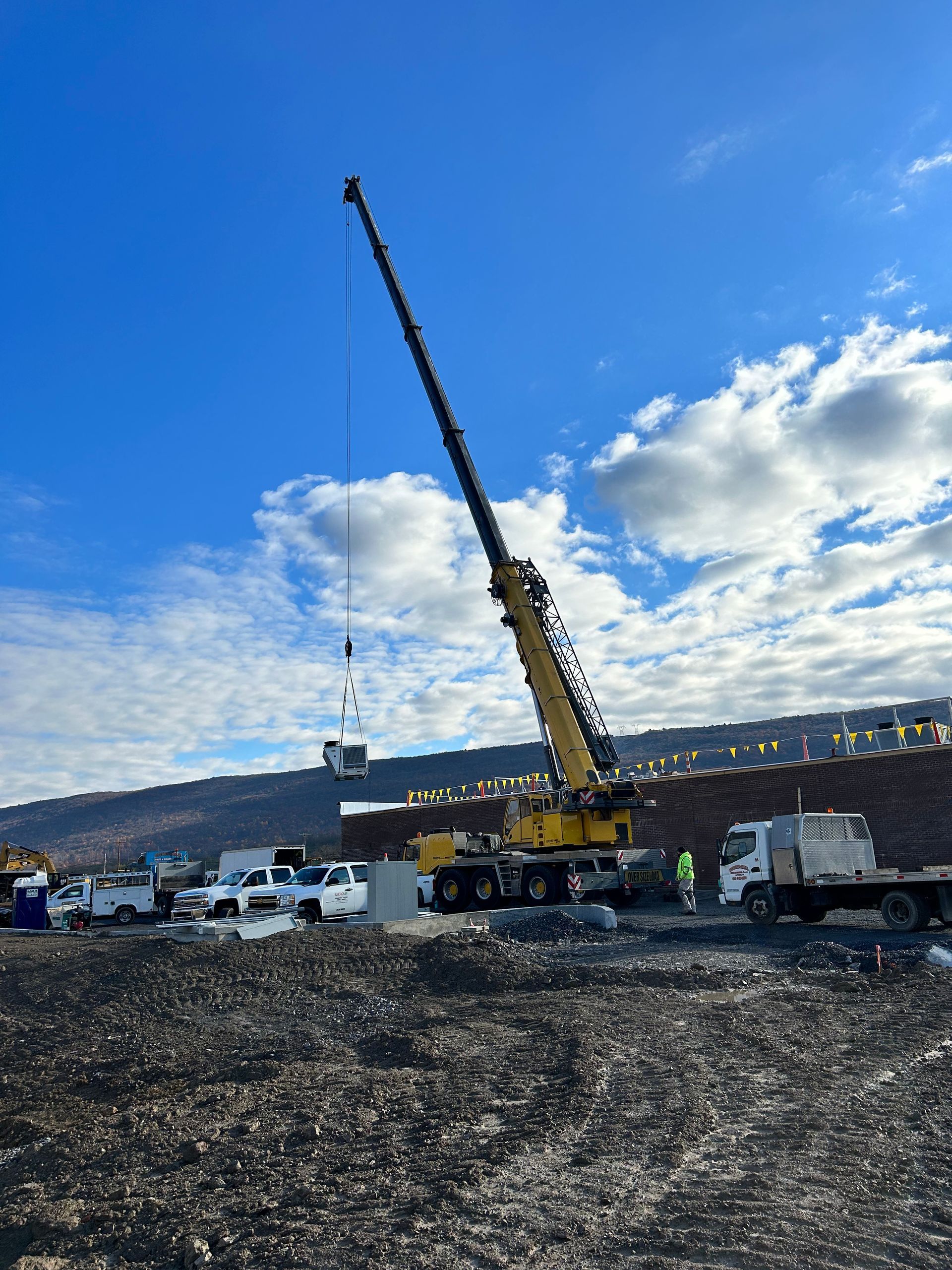 Construction crane raising a long boom over a rocky site with trucks under a blue, cloudy sky