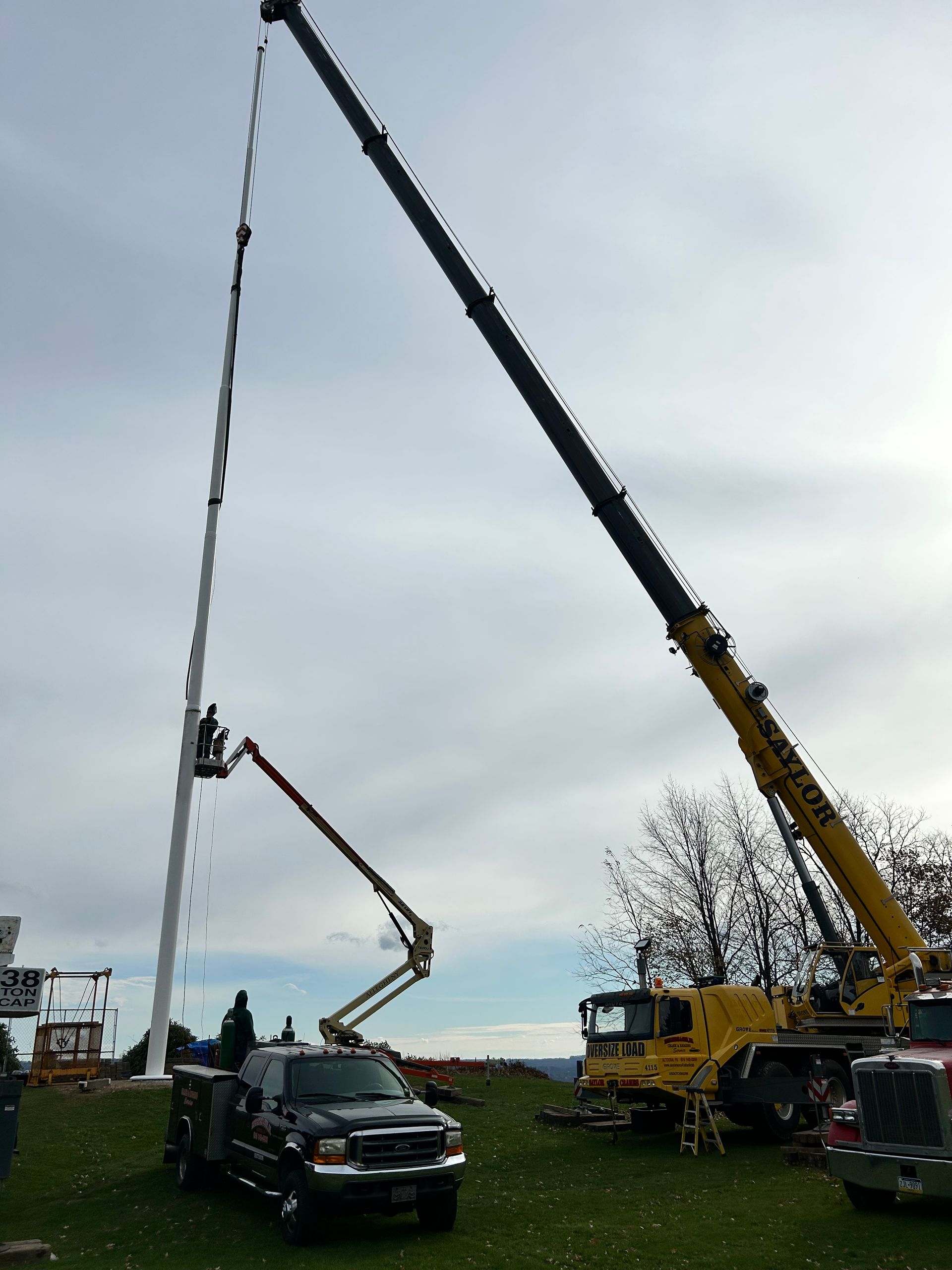 Two cranes lifting a tall, flexible mast beside a black truck in an overcast field