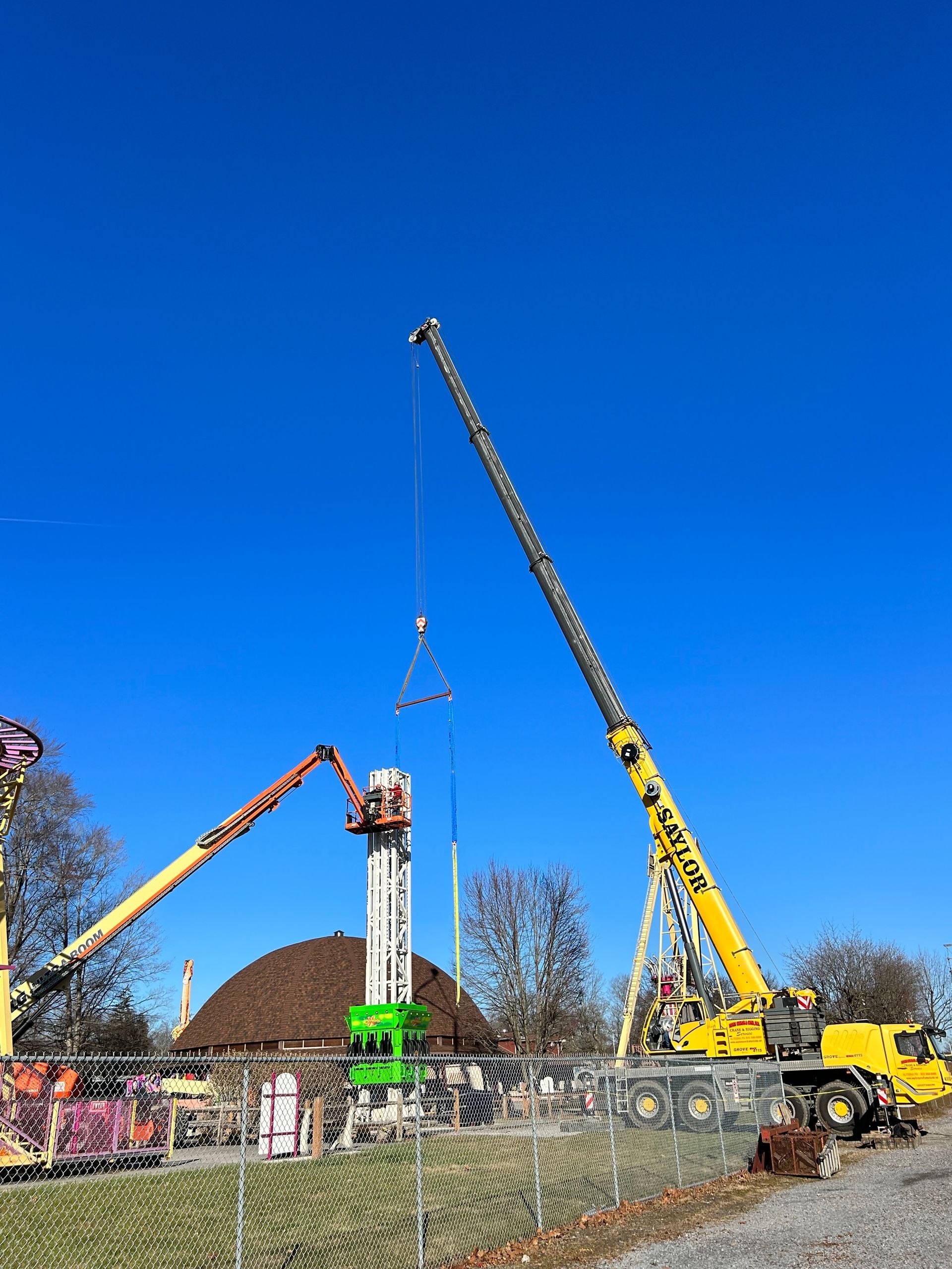 Construction cranes lifting a concrete beam over a fenced site under a clear blue sky