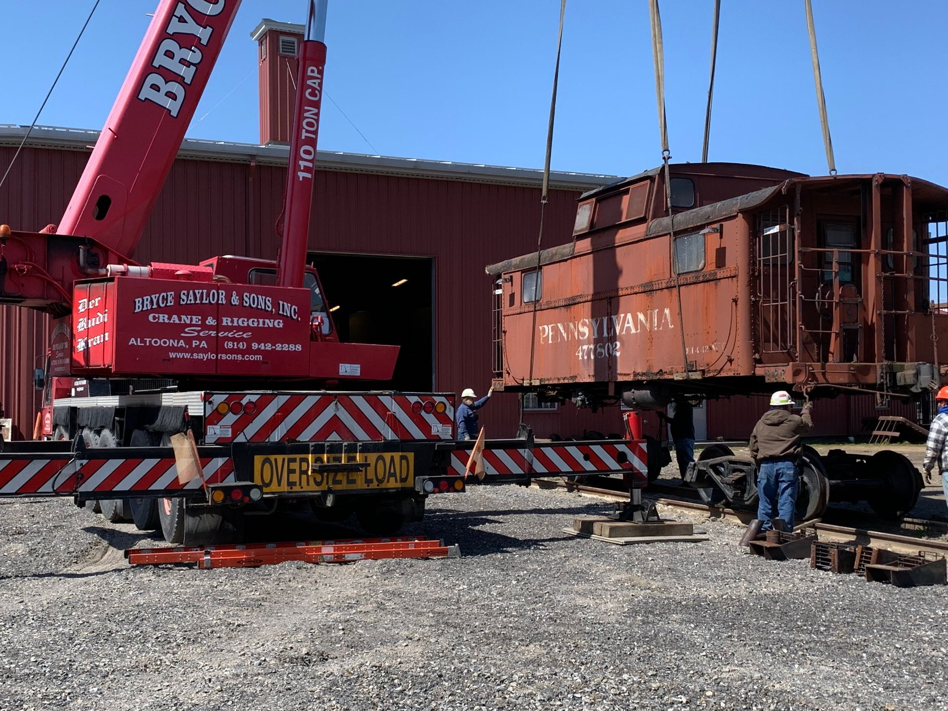 Red crane lifting a rust-colored railcar beside a warehouse, with workers and equipment below.