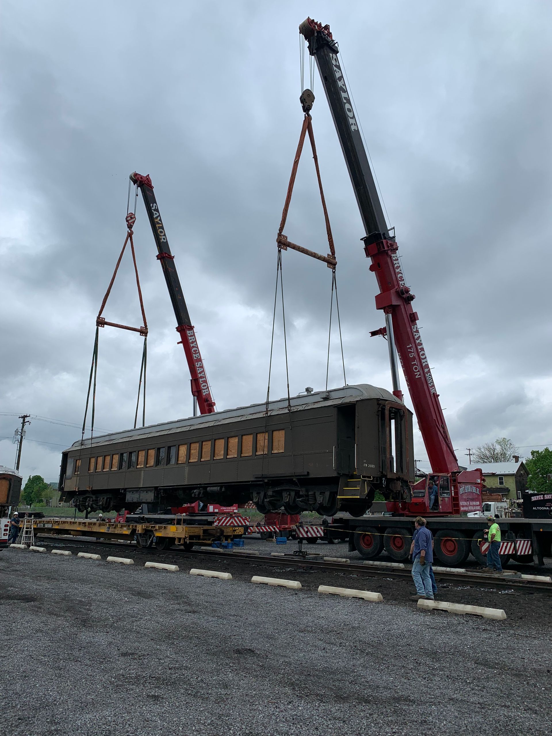 Two cranes lifting a rail car in a rainy industrial yard under cloudy skies