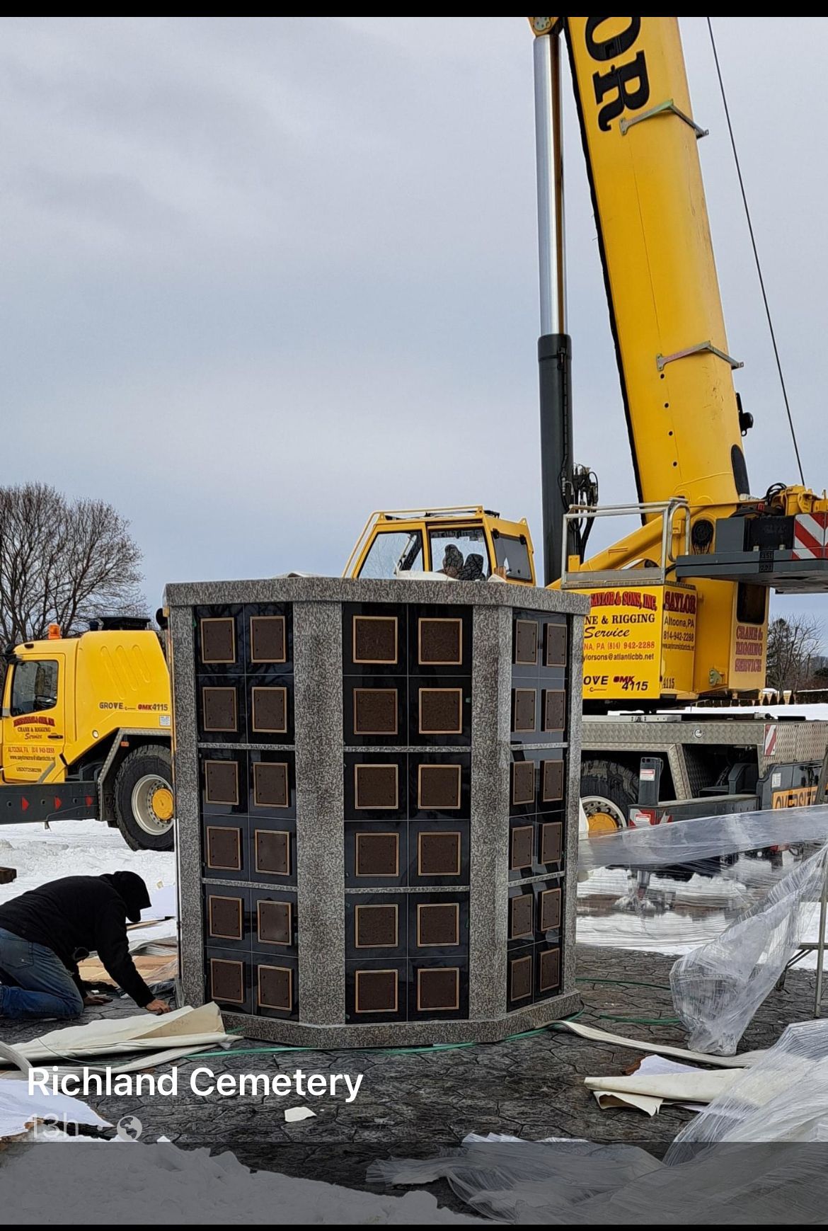 Crane lifting a large stone memorial marker at a cemetery in winter, with workers nearby