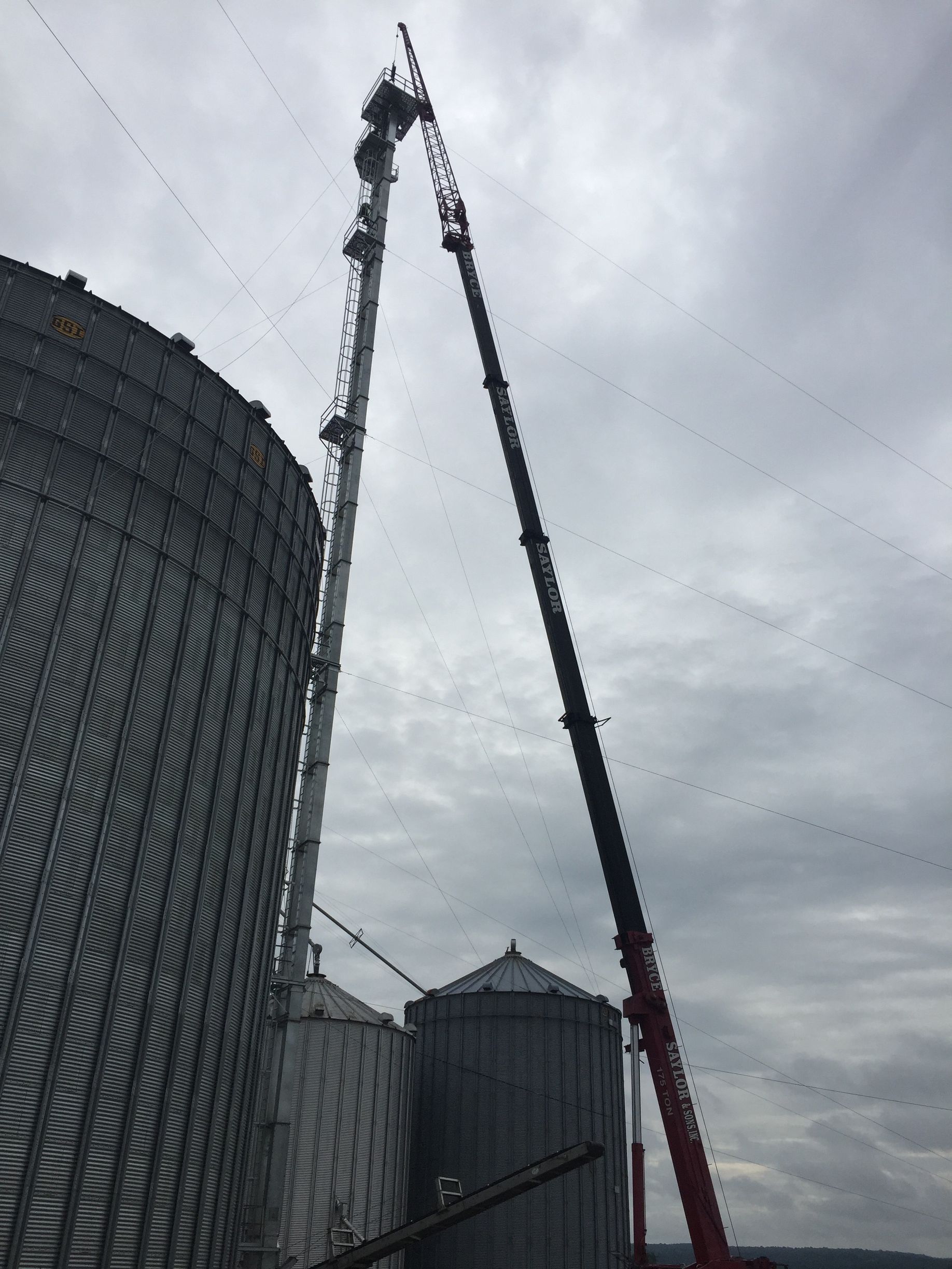 Two cranes lifting a large structure beside silos under a cloudy sky
