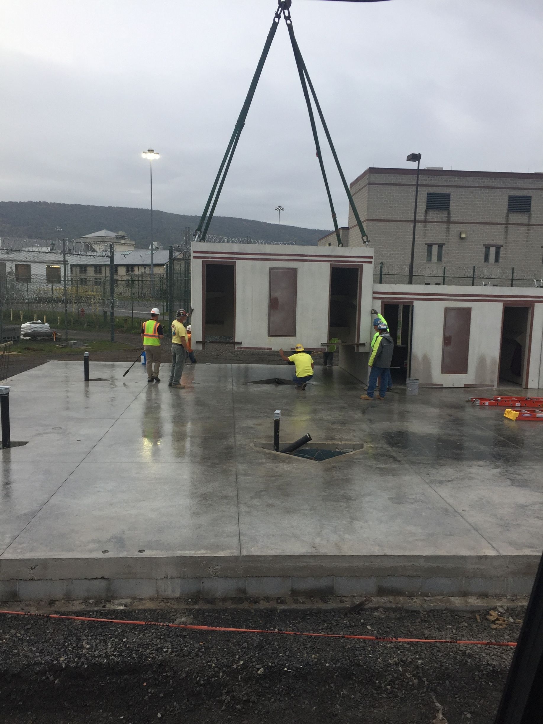 Construction workers hoisting a small concrete structure with a crane on a wet rooftop under cloudy skies