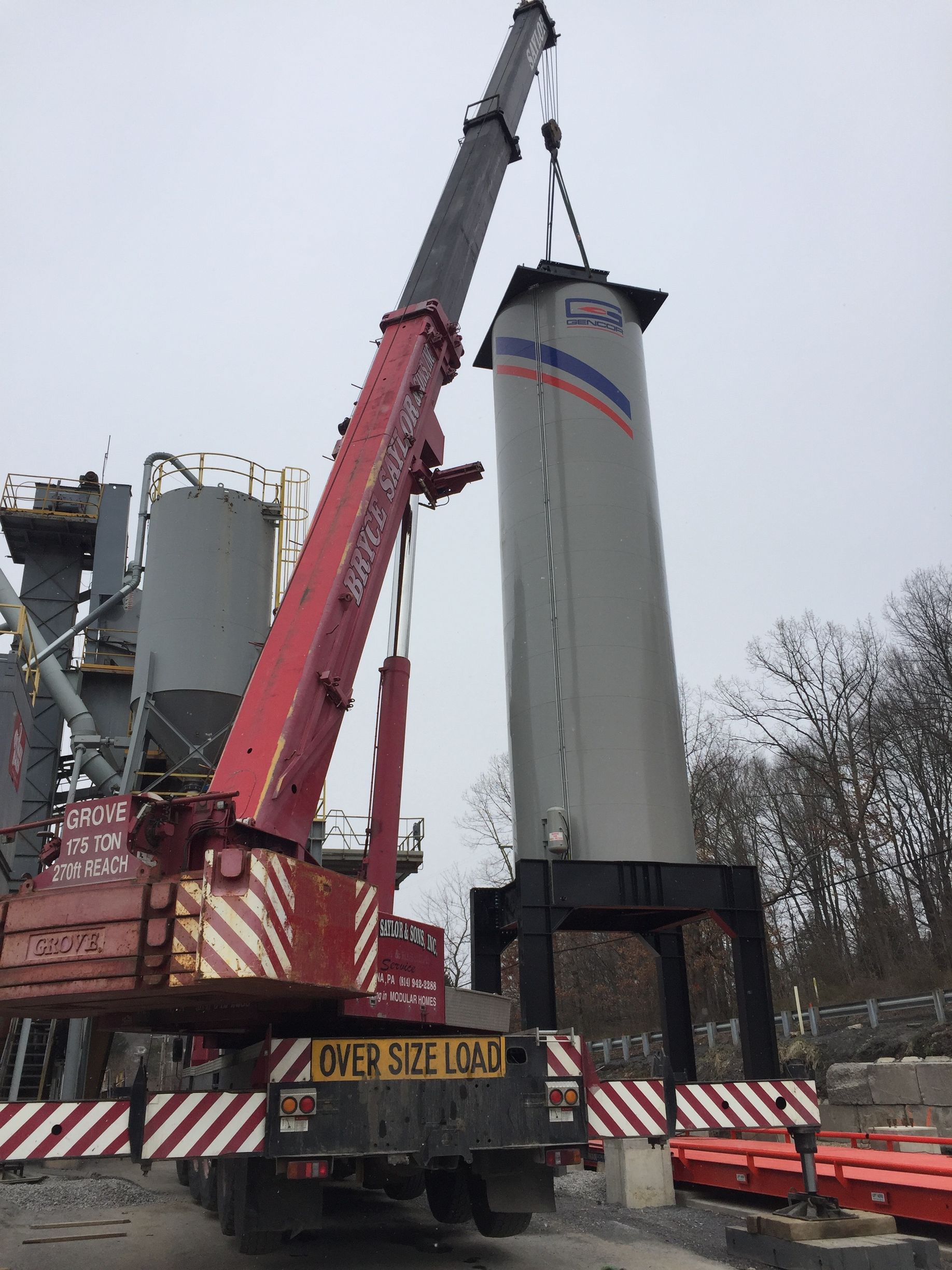Crane lifting a tall white silo section at a construction site, with red support equipment below.