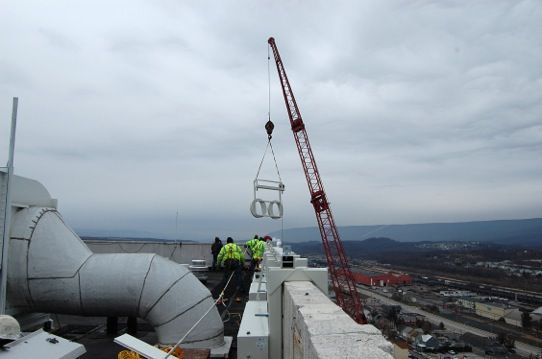 Construction workers on a rooftop lifting a large pipe with a red crane over a cityscape