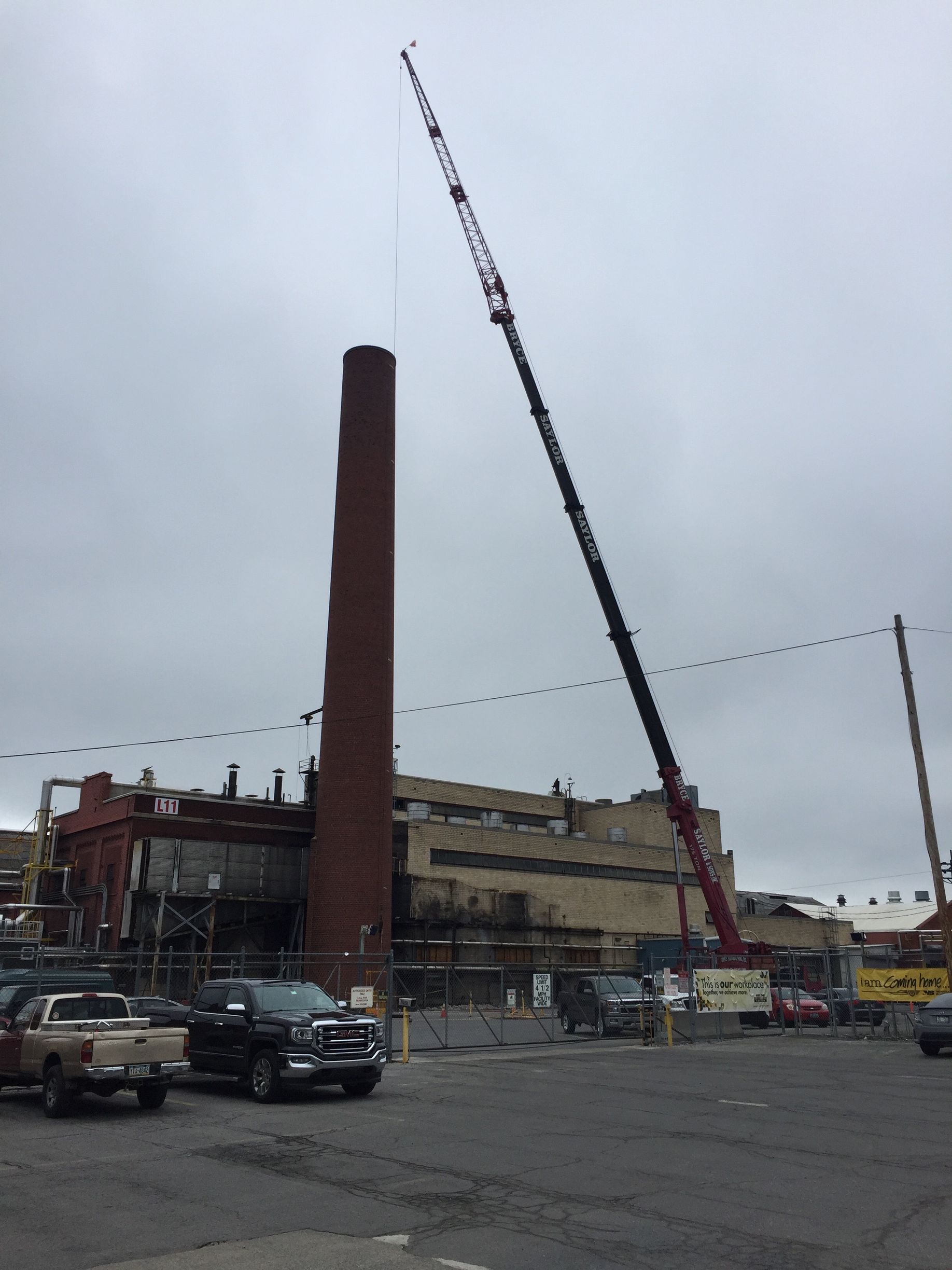 Construction site with a tall crane beside a brick chimney and parked cars under a cloudy sky