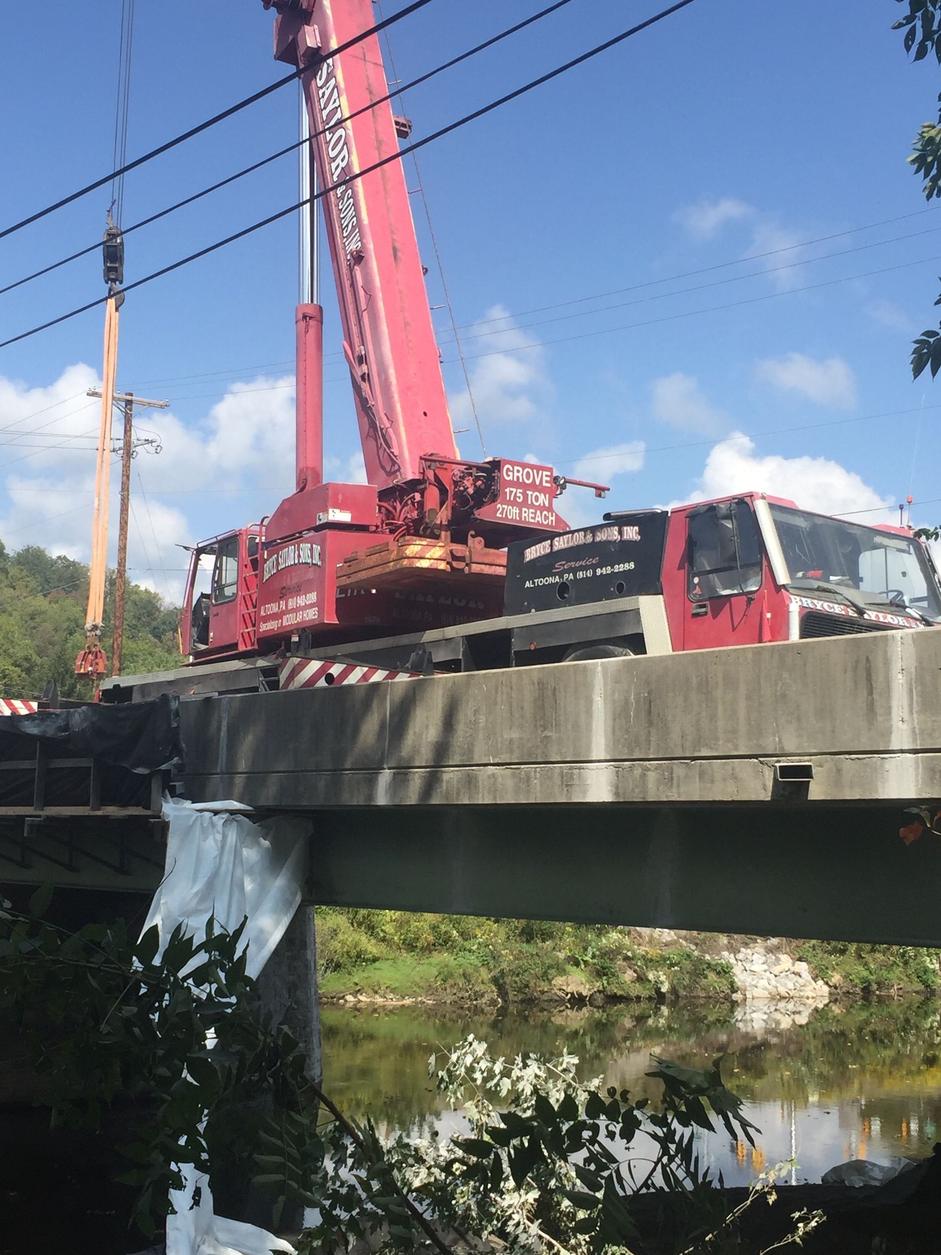 Red crane truck on a bridge over a creek, with utility poles and blue sky nearby.