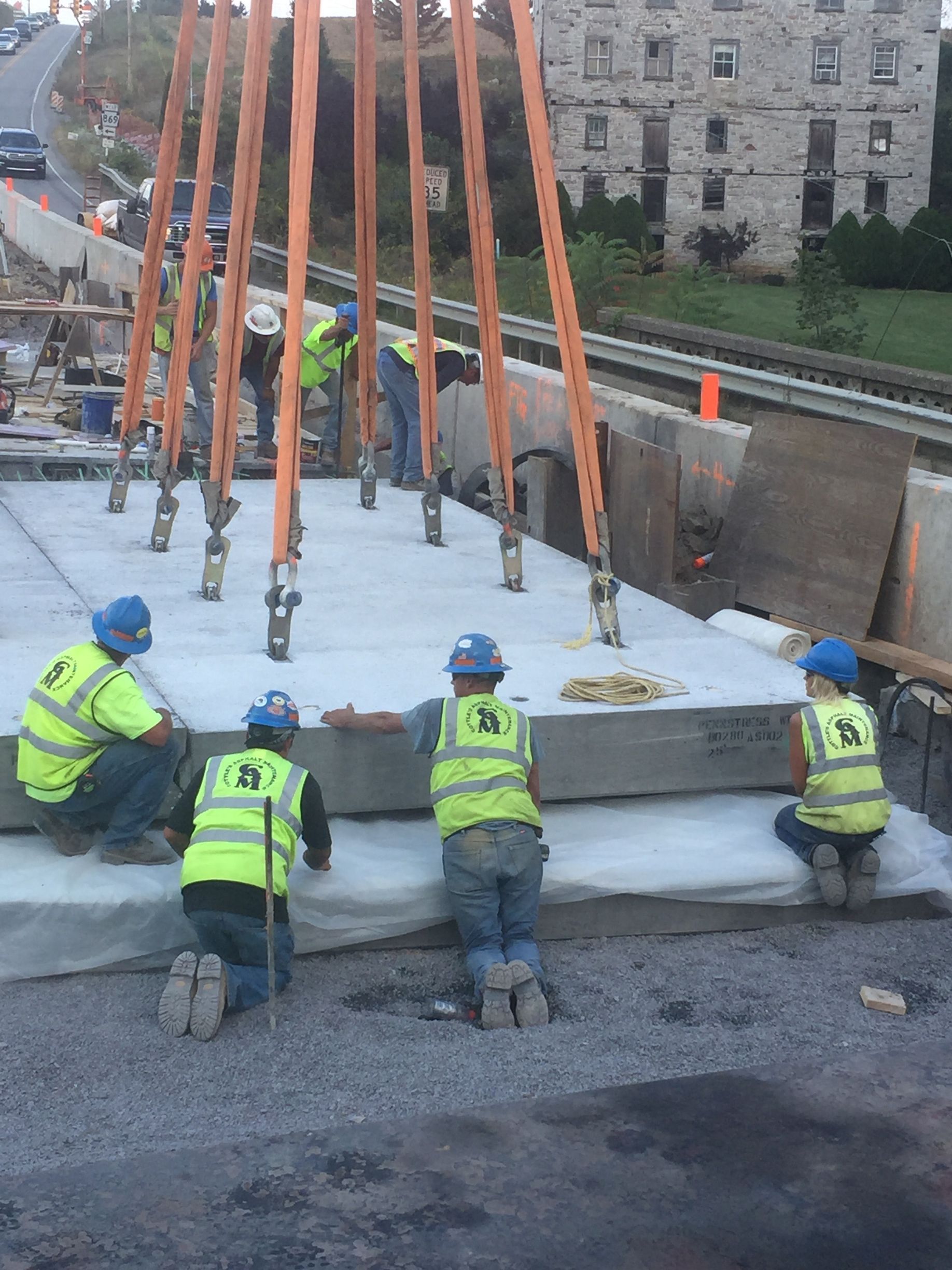 Workers in neon vests secure a large concrete slab at a construction site using orange straps.