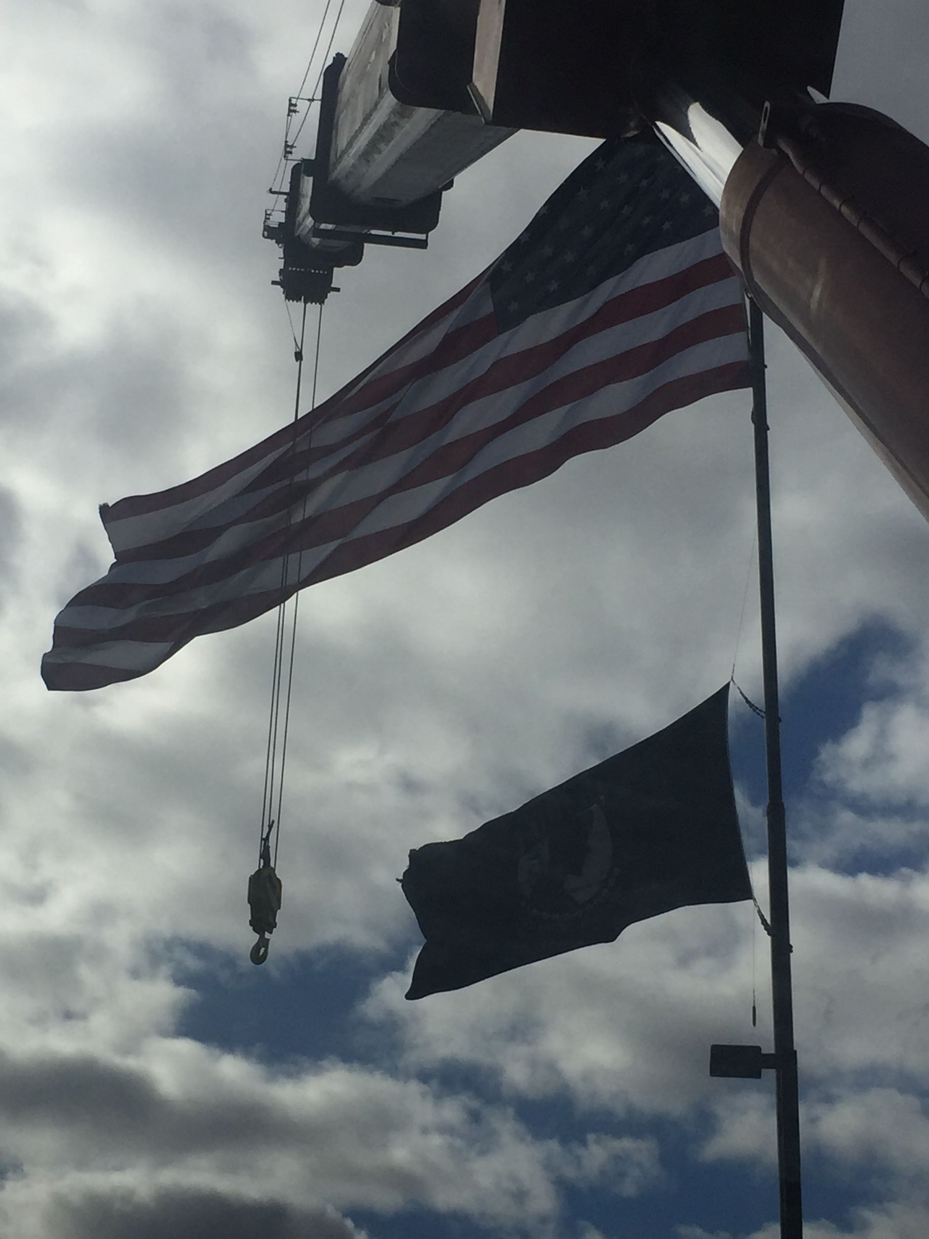 American flag flying on a pole with a dark flag below, silhouetted against a cloudy sky