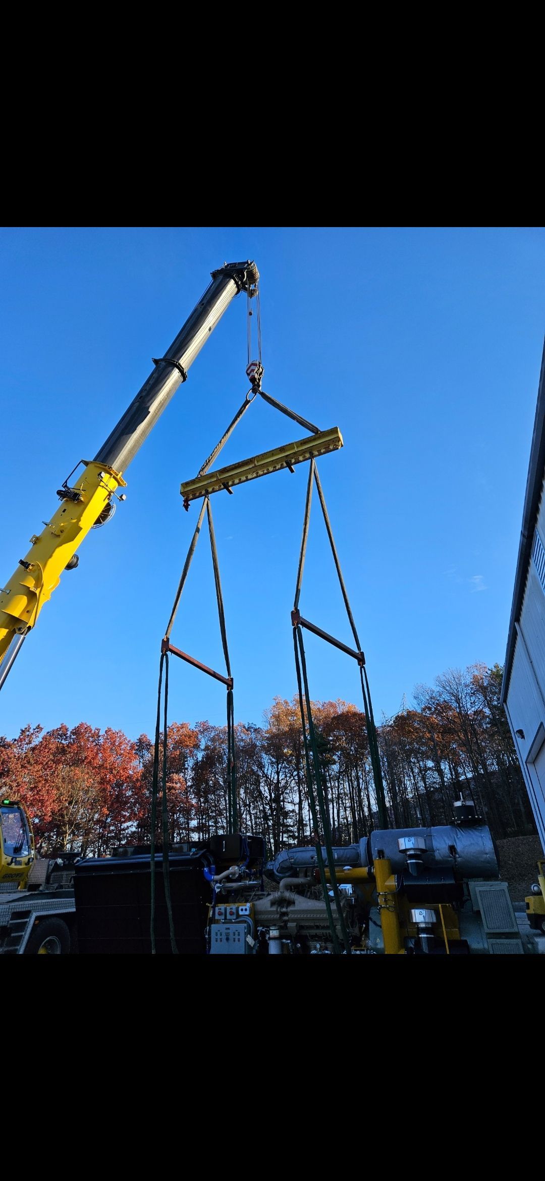Yellow crane lifting a large metal frame against a clear blue sky over trees and equipment