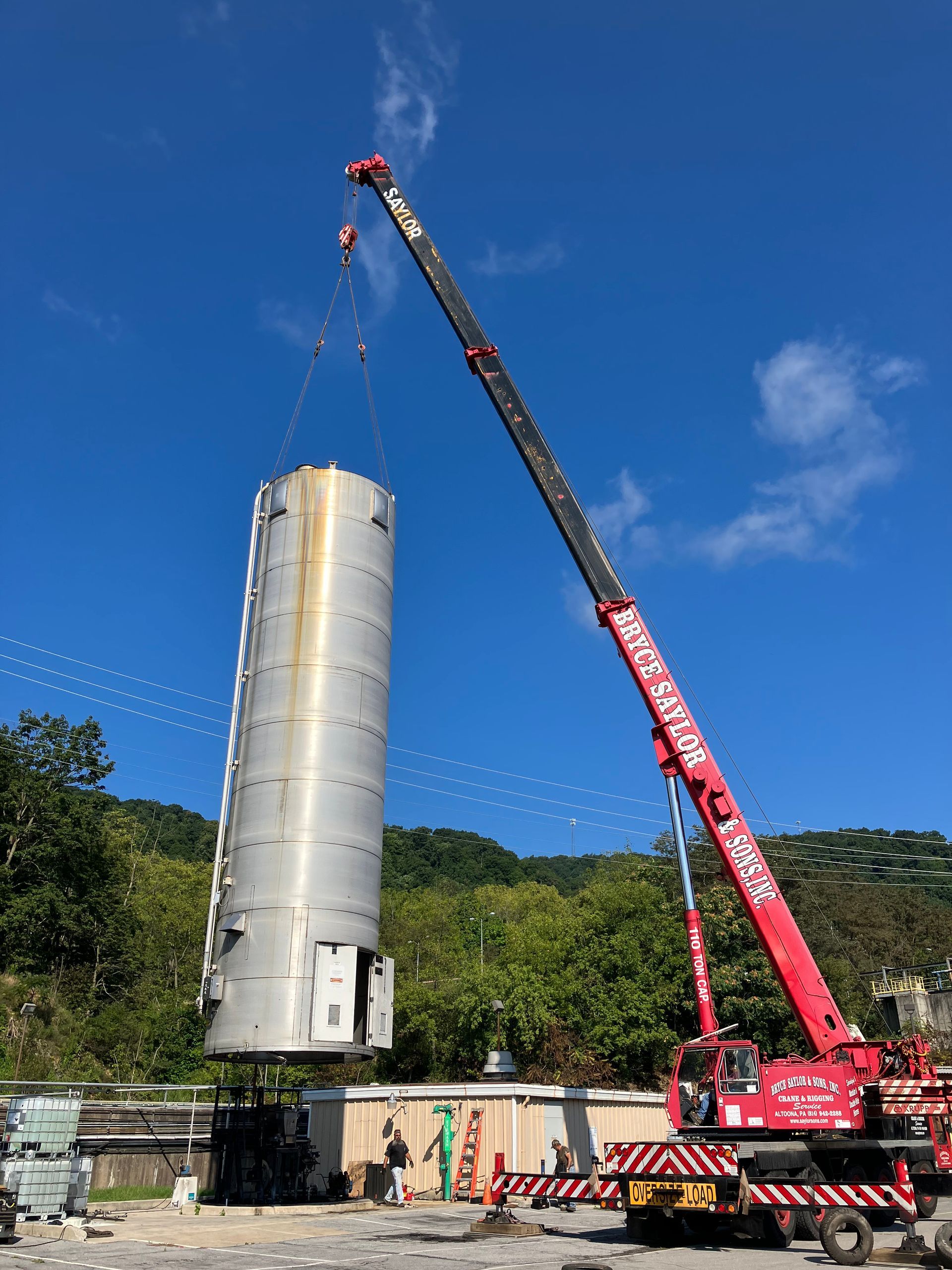 Crane lifting a tall silver cylindrical tank at an industrial site under a blue sky