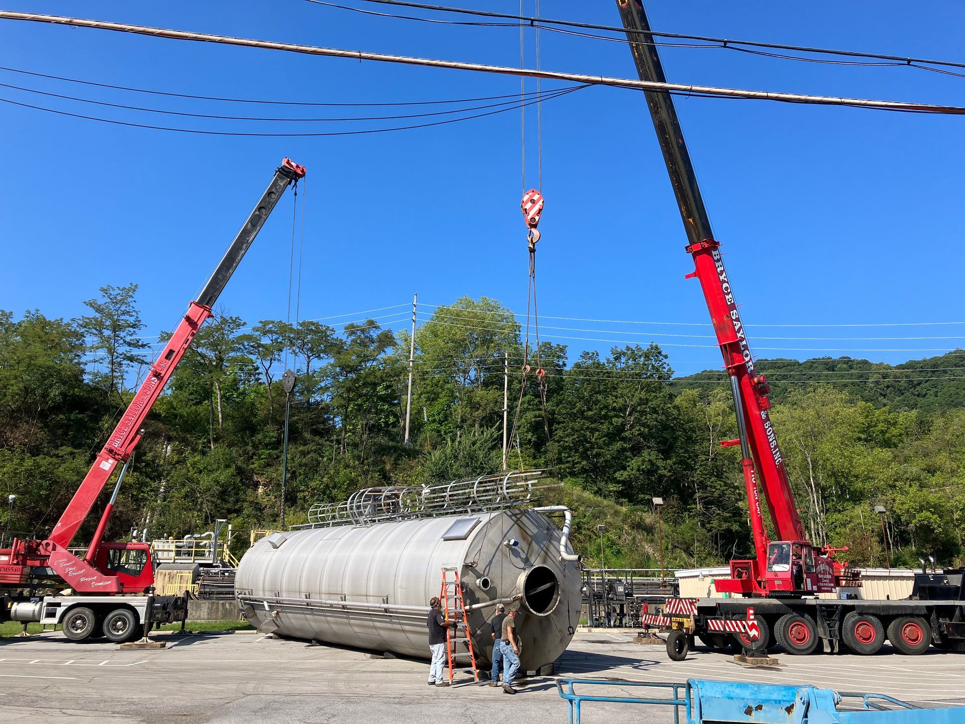 Two red cranes lifting a large cylindrical tank in an open lot under a blue sky