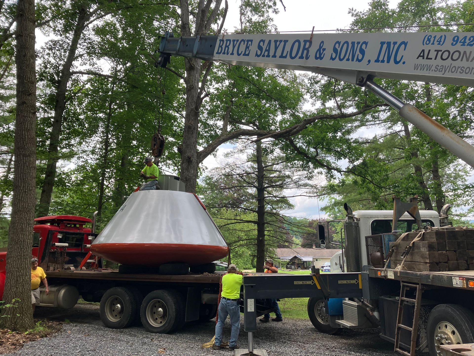 Truck lifting a large conical metal sculpture at a roadside site under trees