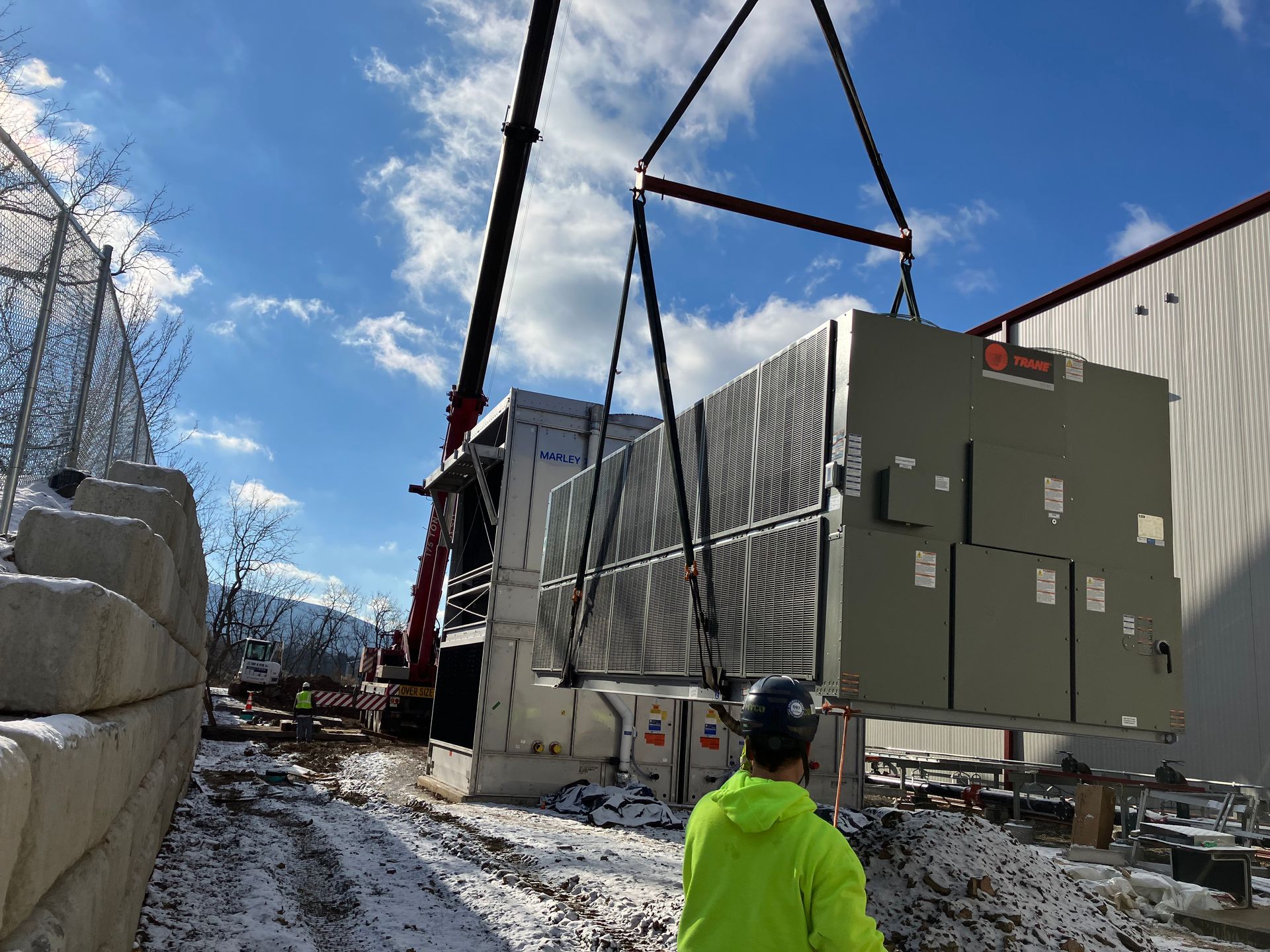 Crane lifting a large green electrical unit beside a building, with a worker in a neon jacket below.