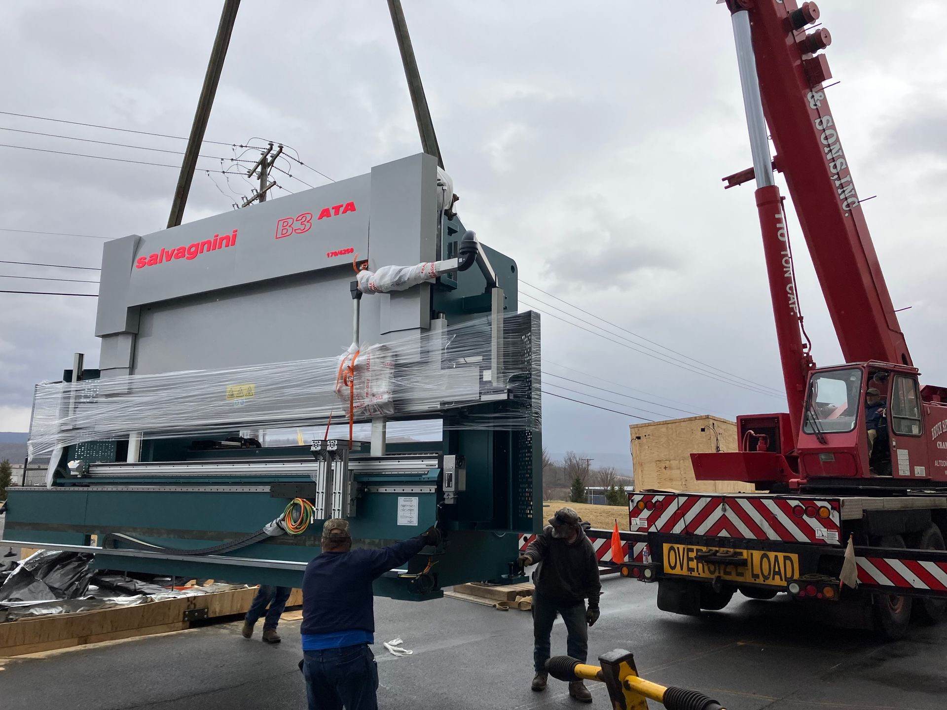 Crane lifting a large wrapped industrial machine beside a truck in a wet lot