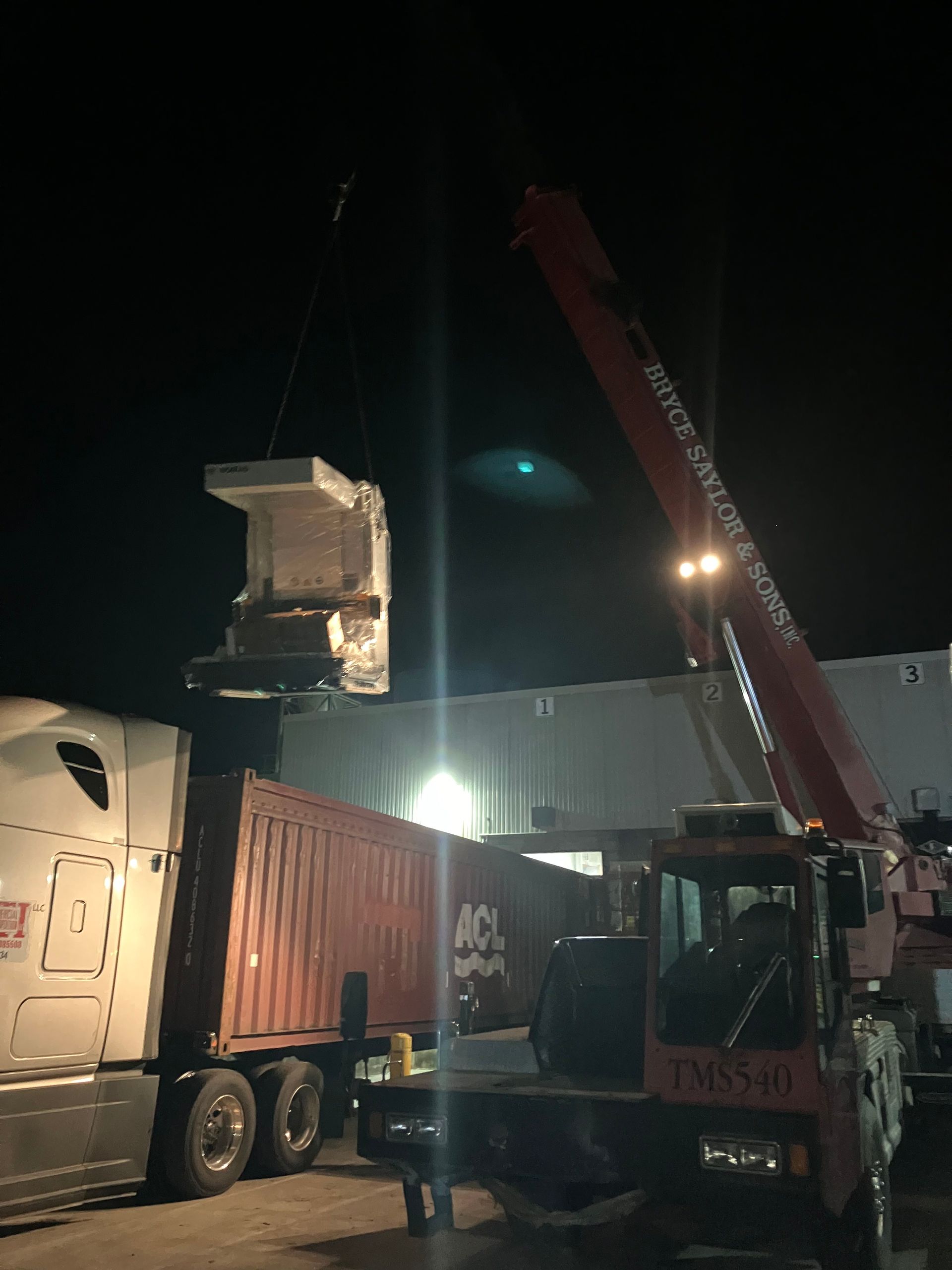 Nighttime crane lifts a large metal container beside a semi-truck in an industrial yard