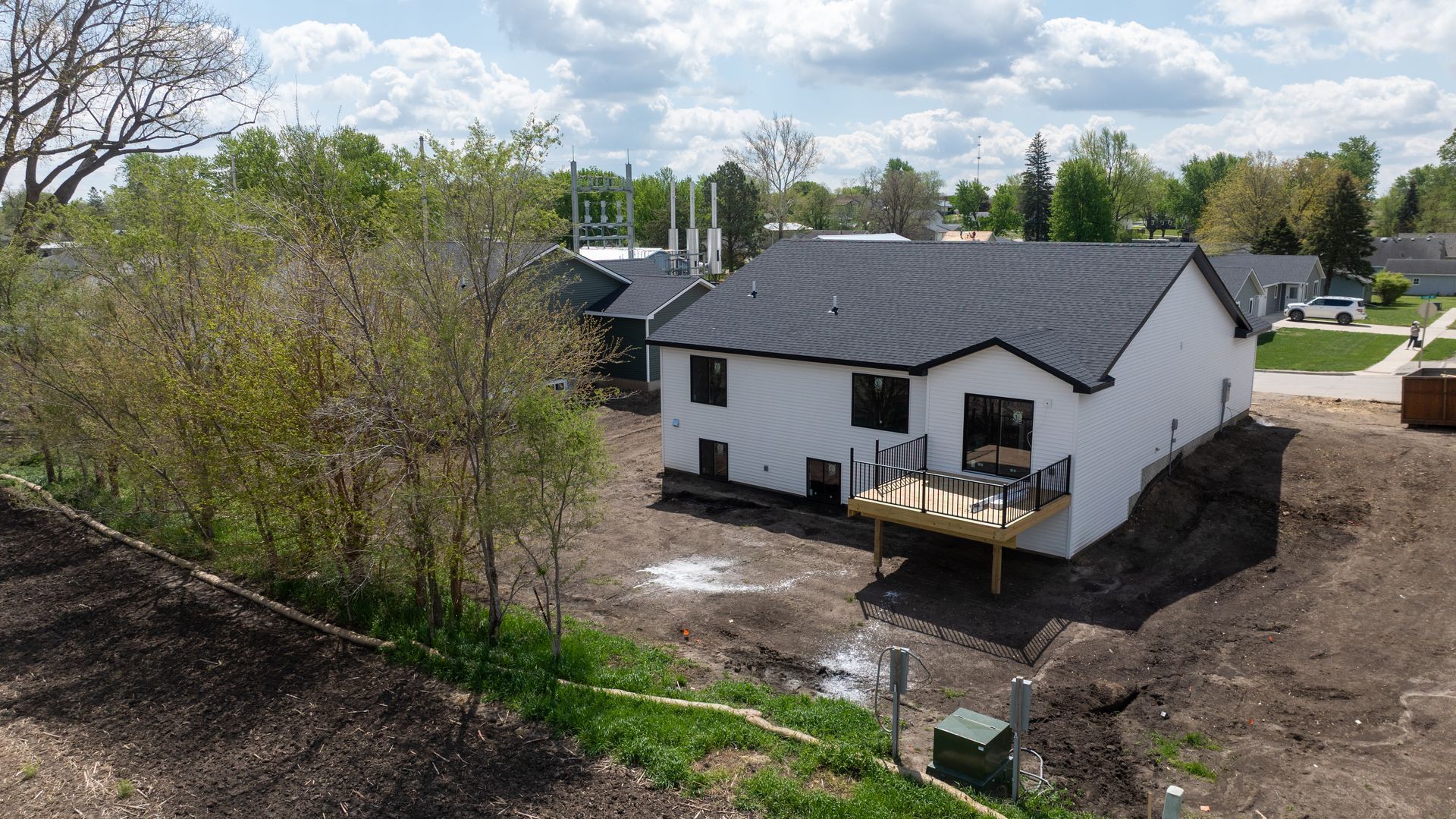 White house with dark roof and small deck on a muddy lot, viewed from a hillside under a partly cloudy sky