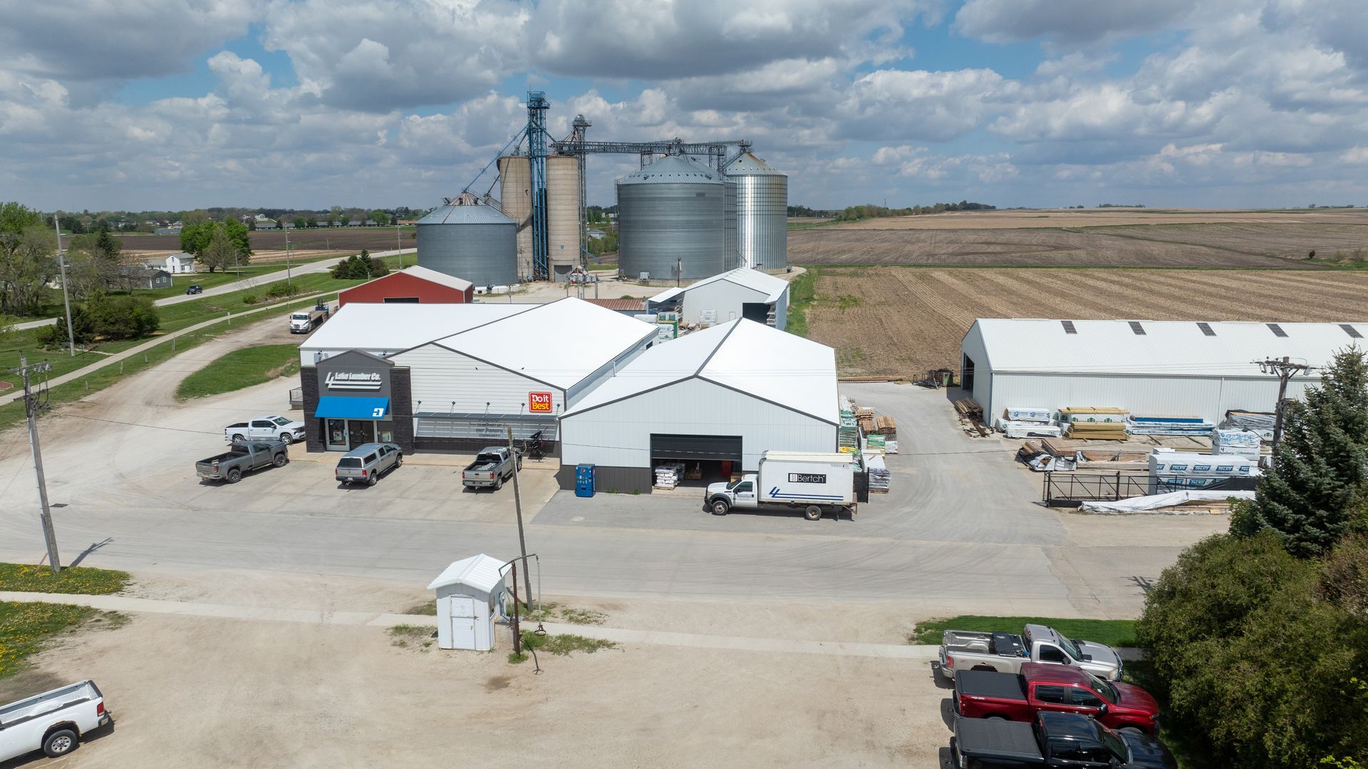 Industrial farm facility with grain silos, white buildings, and parked trucks beside open fields