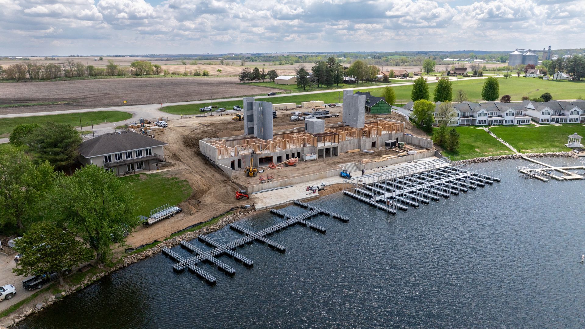 Aerial view of a lakeside marina construction site with docks, buildings, and parked boats.