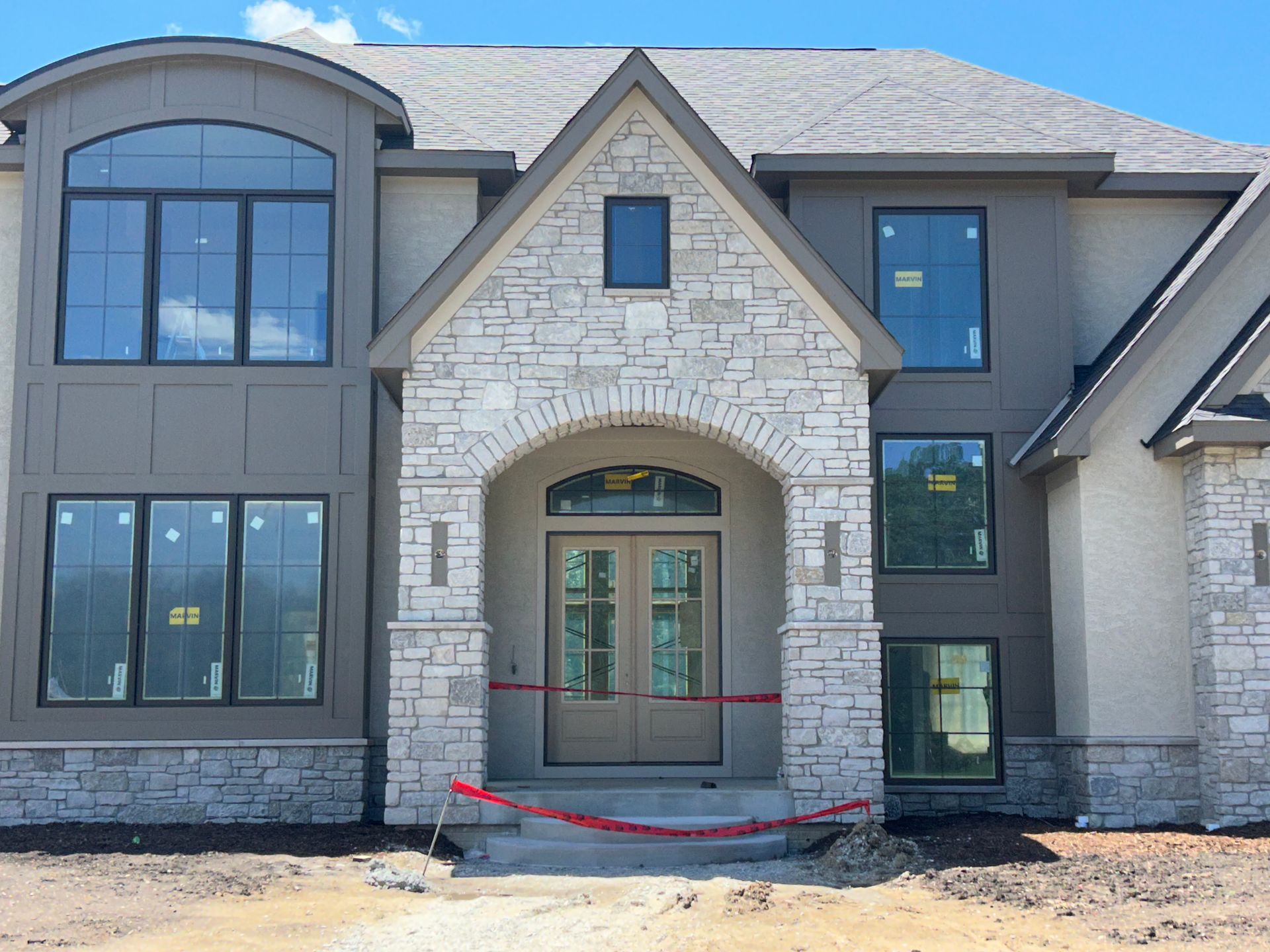 Front view of a modern two-story house with stone facade, large windows, and arched entryway under construction.