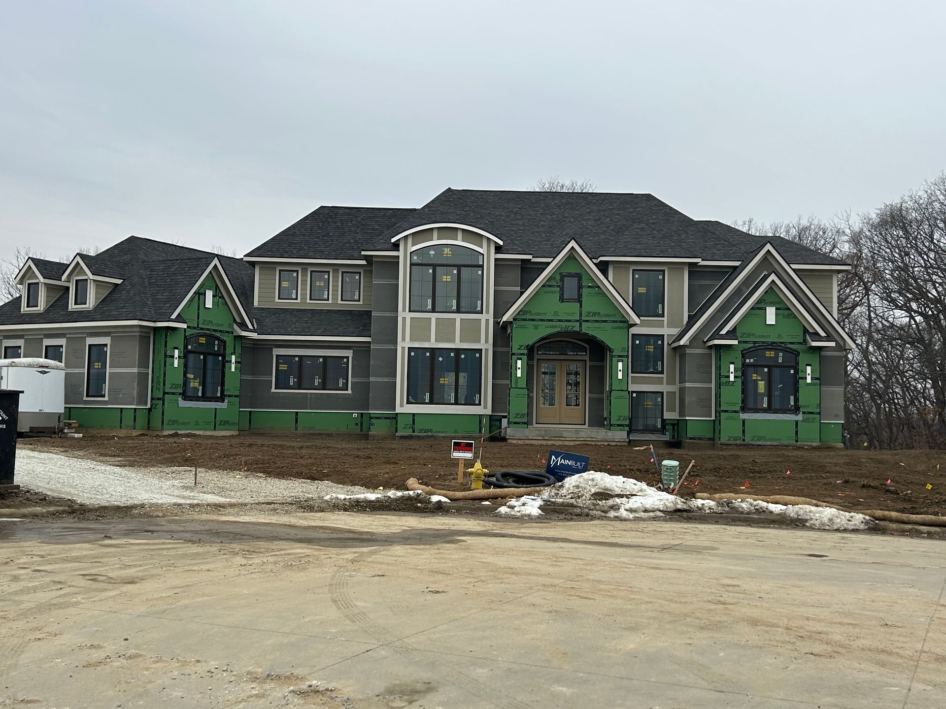 Large house under construction with green and gray siding, snow on the ground, and a cloudy sky.