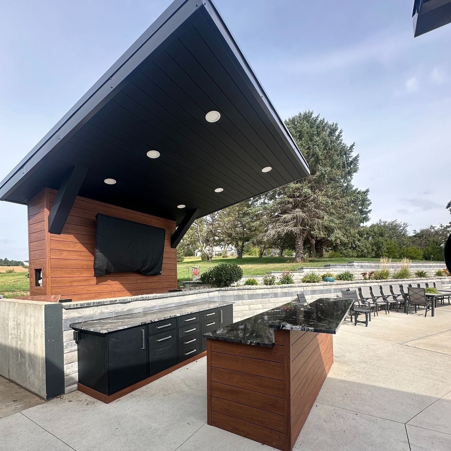 Modern outdoor kitchen and entertainment space with a black roof, wood-paneled walls, built-in television, and stone island.