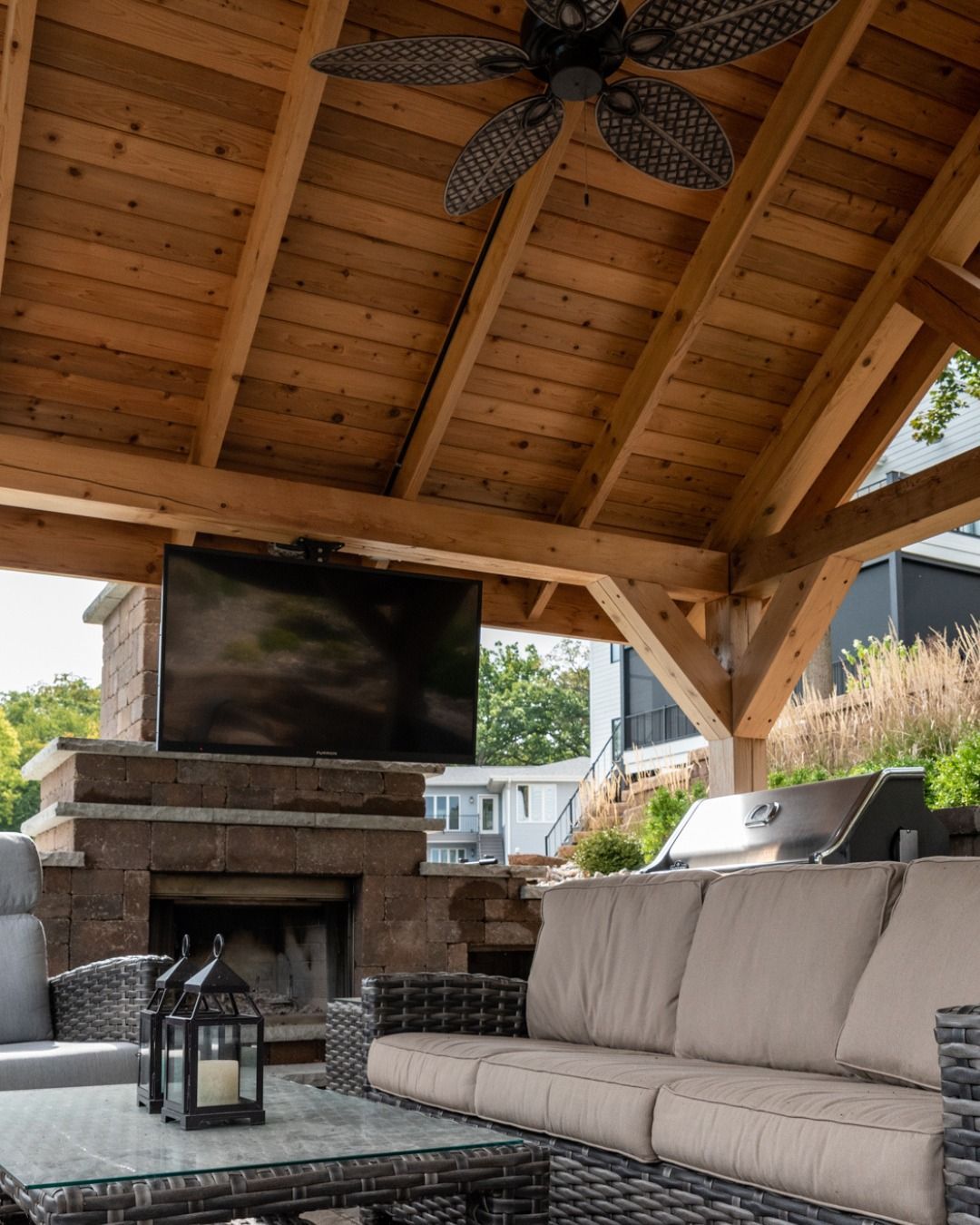 Outdoor covered patio with a fireplace, wall-mounted television, wicker sofa, and a ceiling fan under a wood roof.