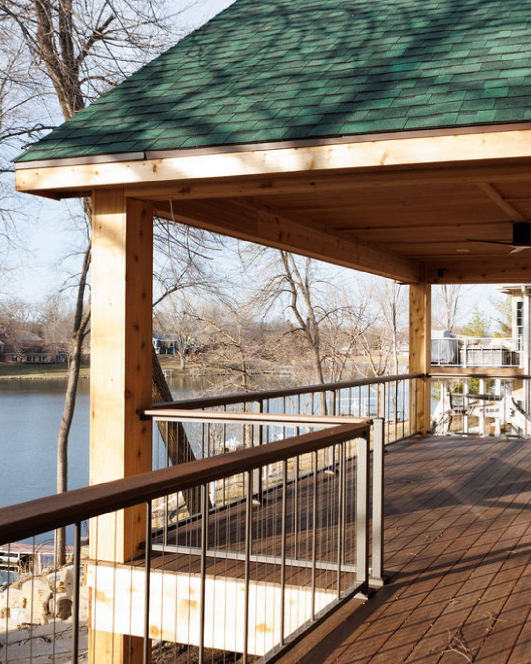 Wooden deck overlooking a lake, with green-roofed pergola and railing in a wooded setting