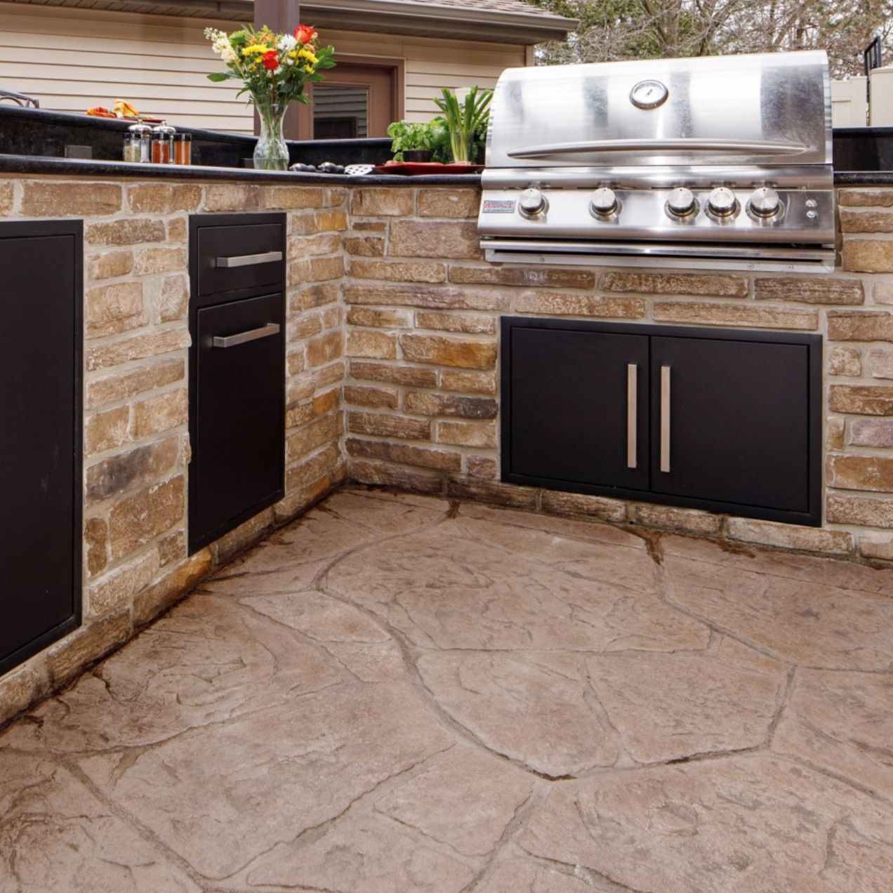 Outdoor built-in stone kitchen with stainless steel grill, black cabinets, and countertop planters