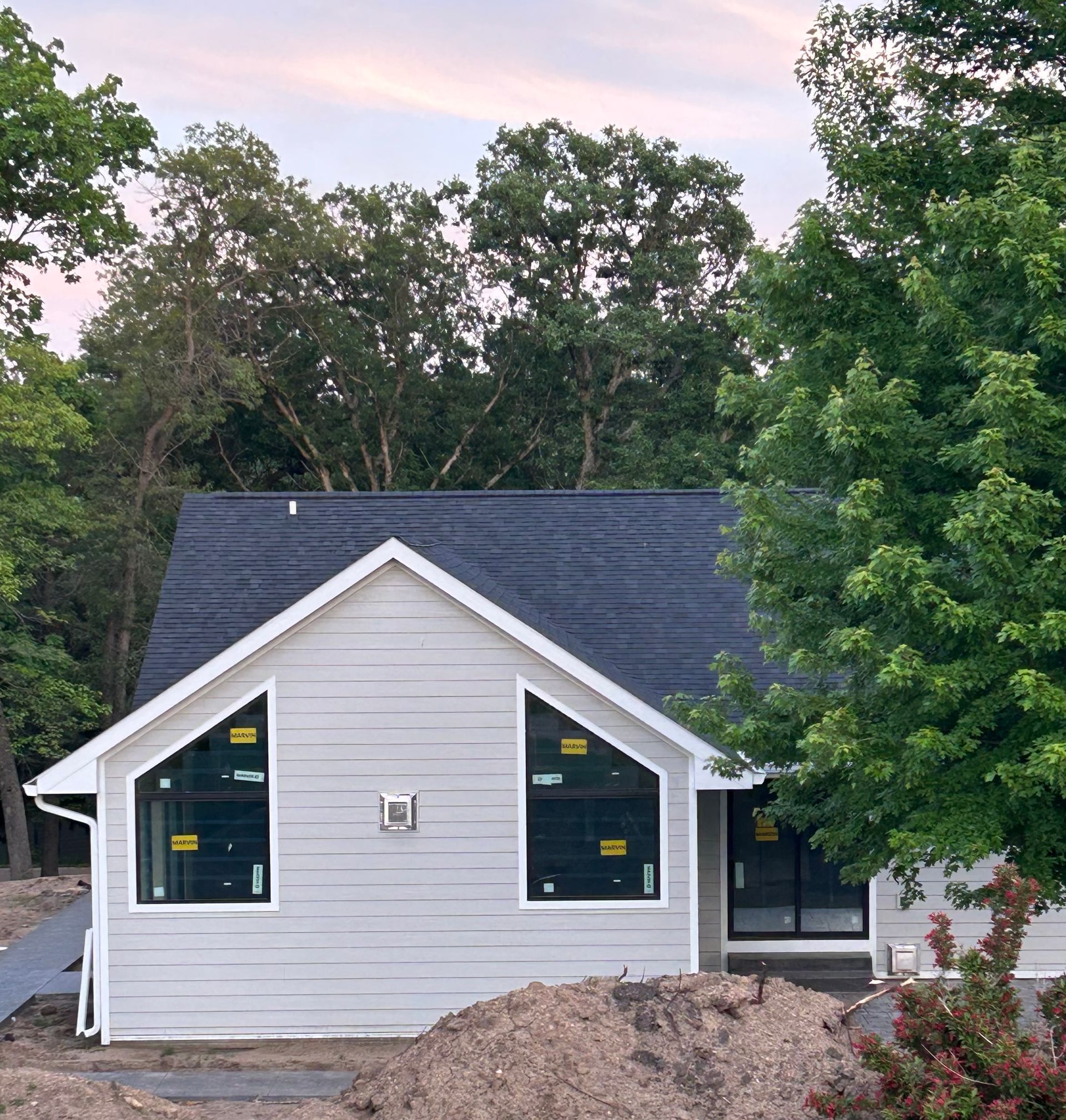 Small gray house with black roof and large windows, surrounded by trees and dirt in the foreground.