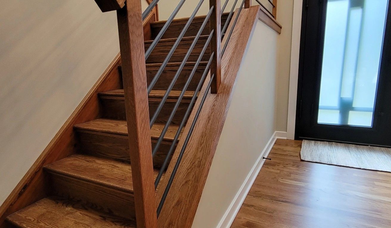 Wooden staircase with horizontal metal railing in a home entry with light-colored walls and a black-framed front door.