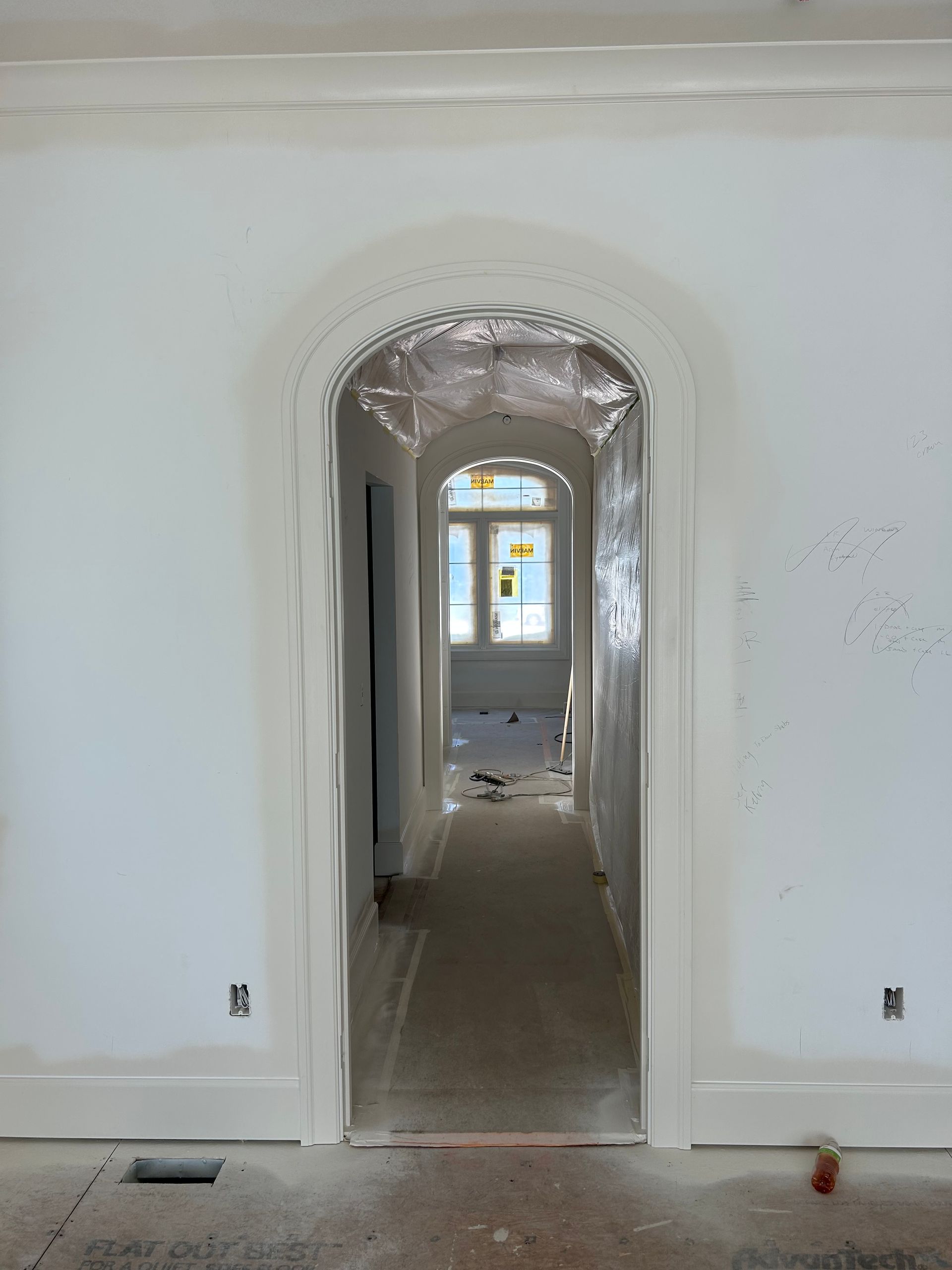 White hallway with arched doorway, plastic-covered opening, and a window at the far end during renovation.