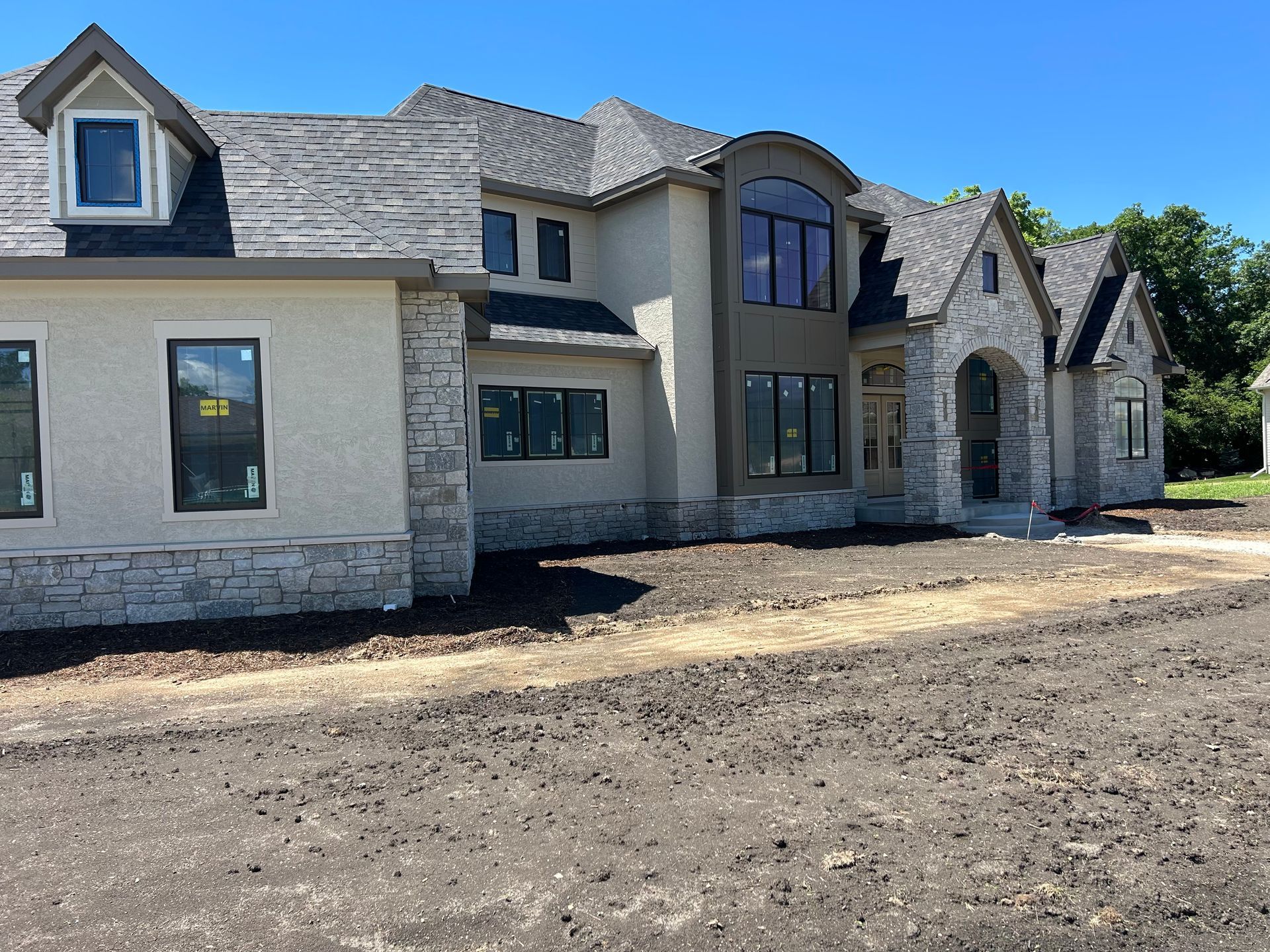 Stone-and-stucco house under construction with multiple rooflines and bare dirt foreground
