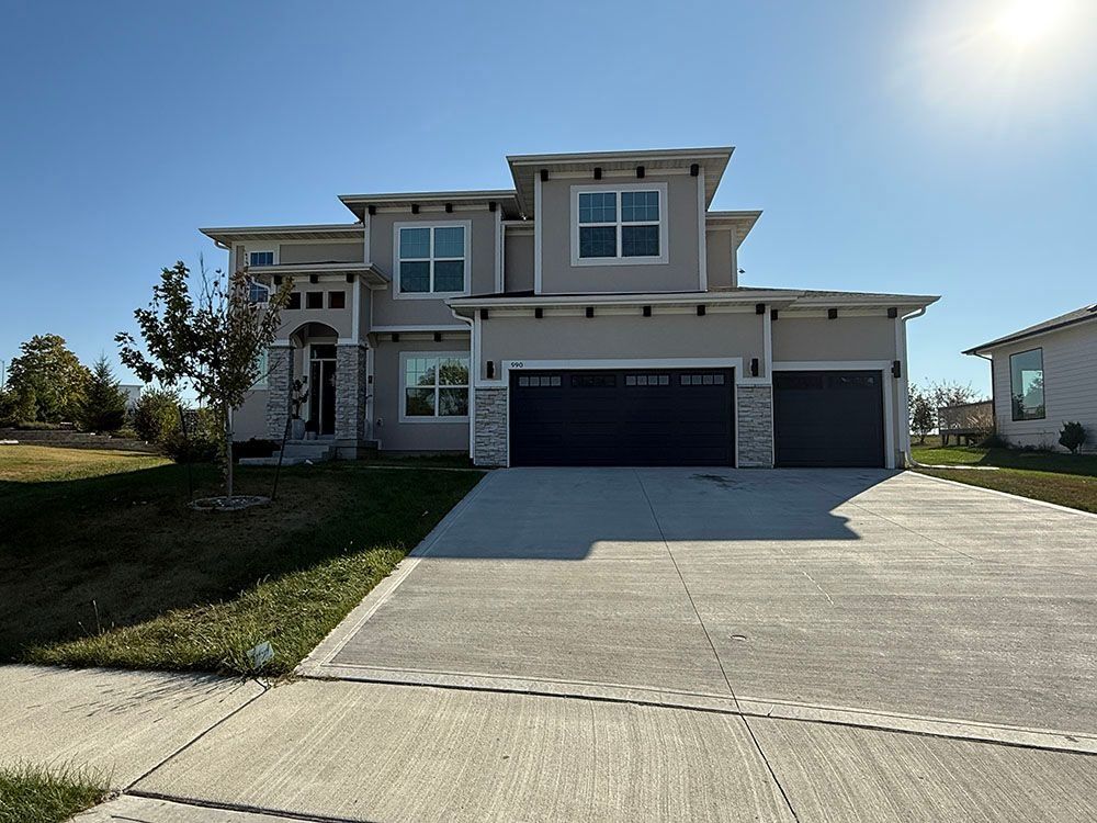 Modern two-story house with gray stucco exterior, stone accents, black garage doors, and a concrete driveway on a sunny day.