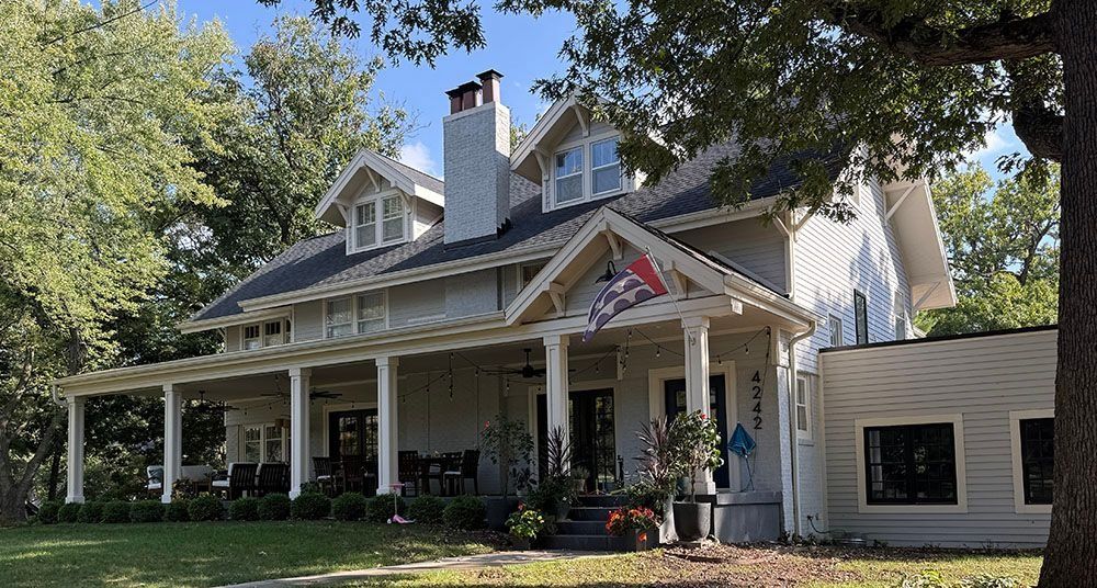 Gray two-story house with a long front porch and a flag; surrounded by trees.