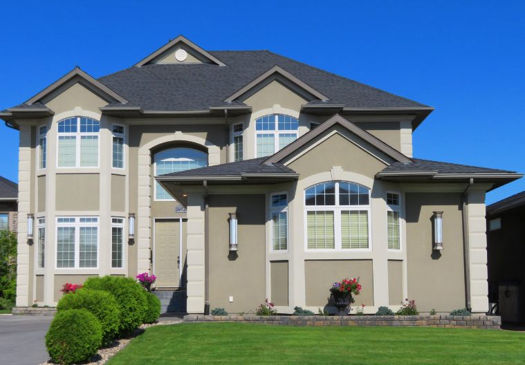 Two-story beige house with dark roof and trim, several windows, and manicured lawn. Blue sky background.