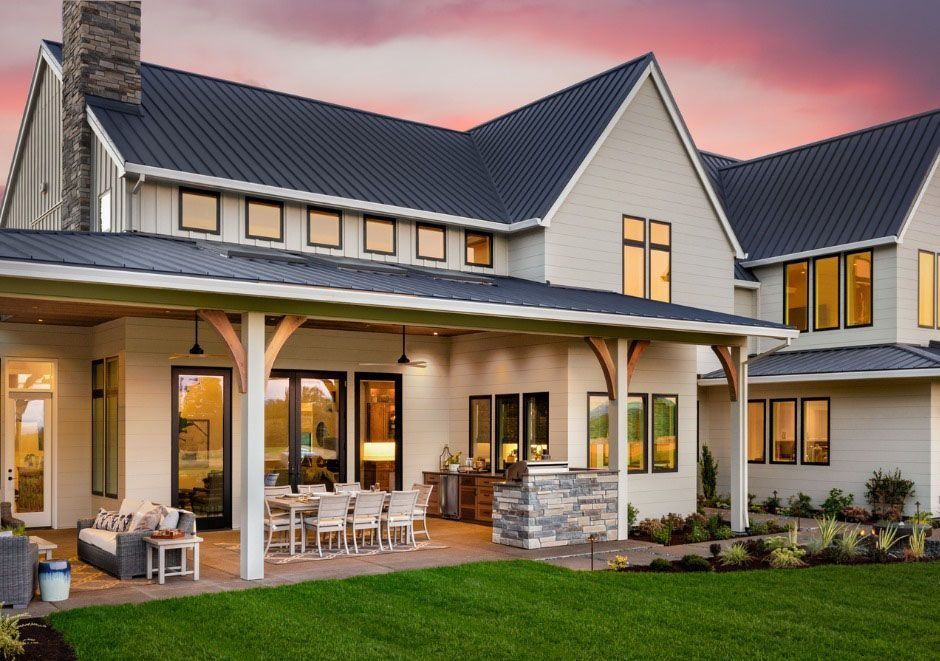 A modern white house with a dark roof and large windows, a covered patio with dining, and a green lawn.